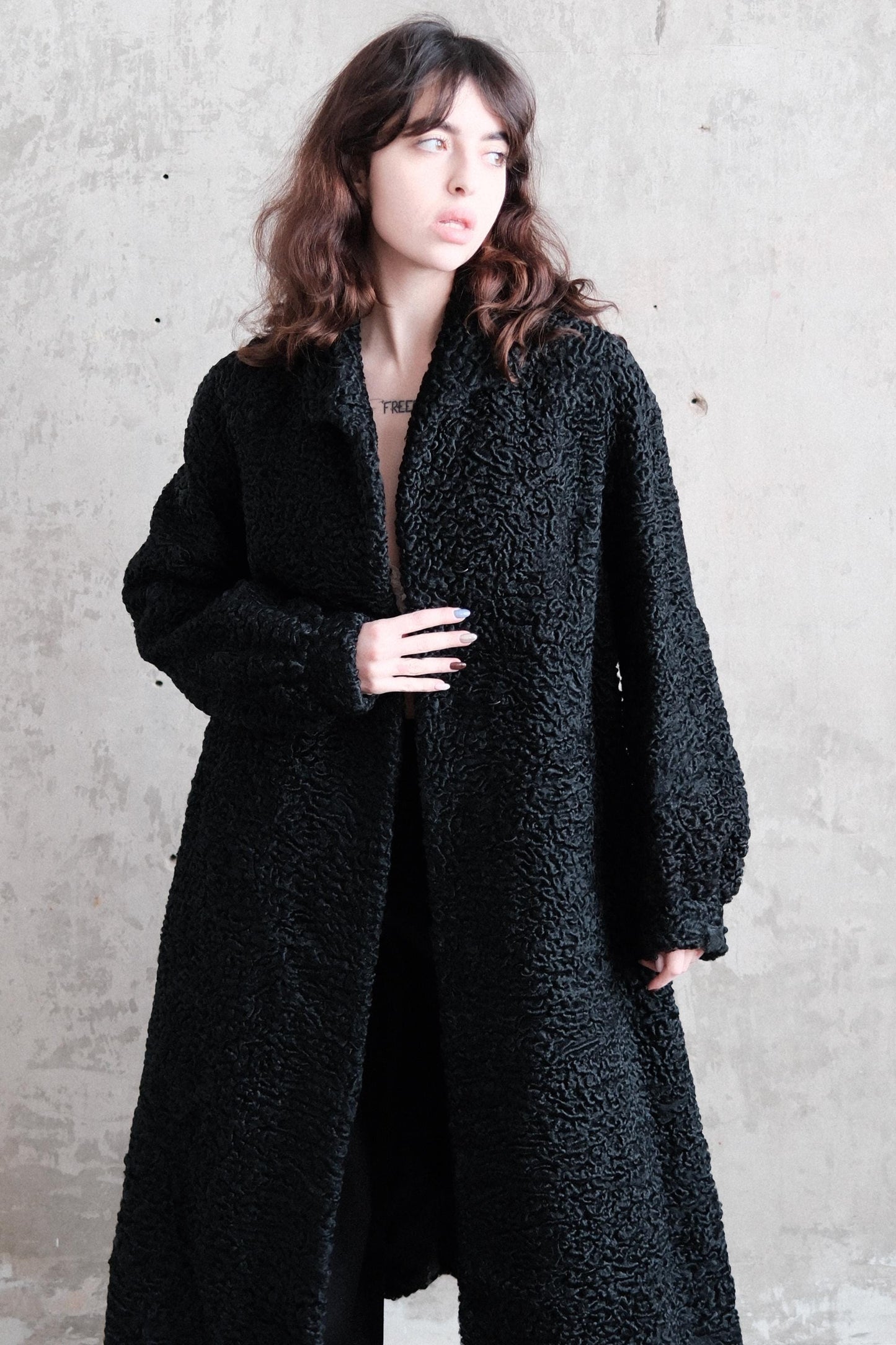 A woman wearing a long black coat is posing in a relaxed manner against a plain background.
