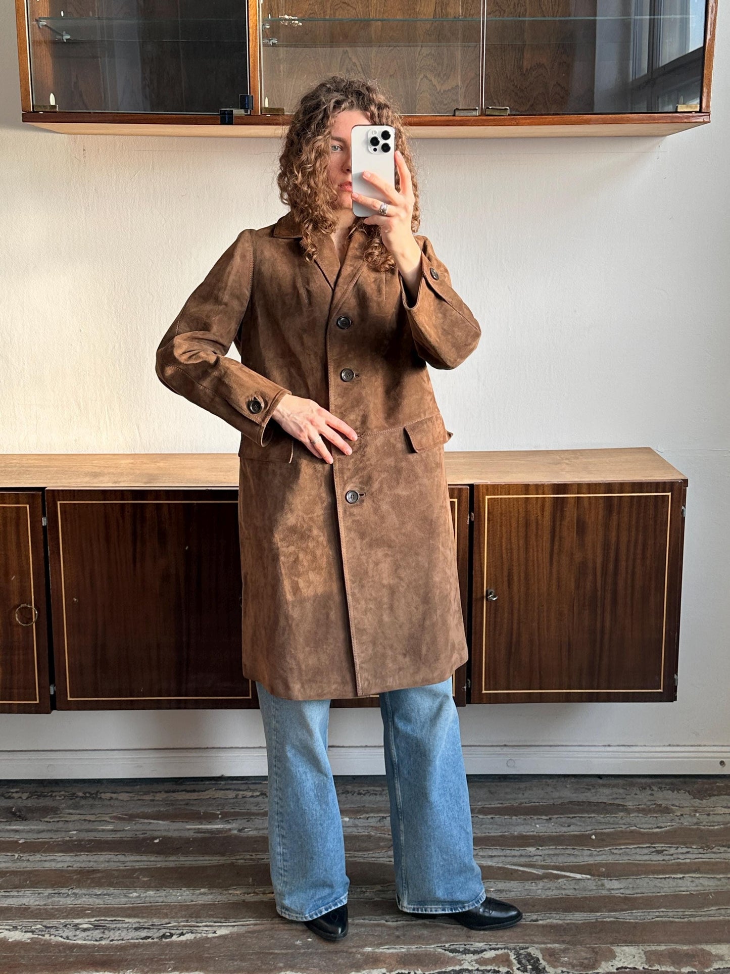 A person is taking a selfie with a brown coat and blue jeans, standing in front of a wooden cabinet.