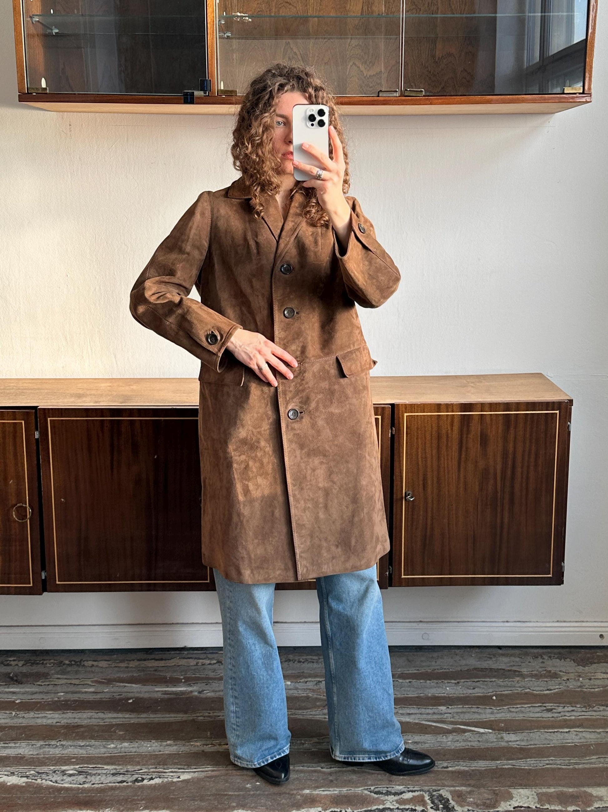 A person is taking a selfie with a brown coat and blue jeans, standing in front of a wooden cabinet.