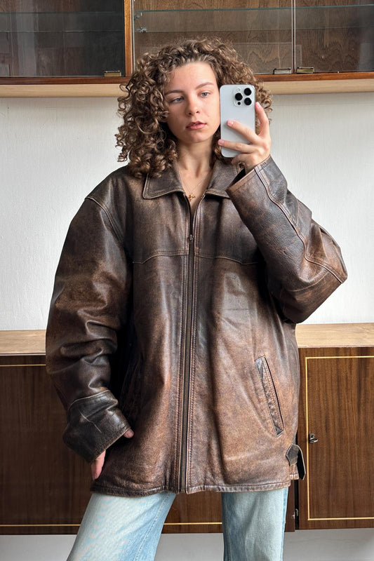 A woman with curly hair is taking a selfie with a smartphone, wearing a brown leather jacket.