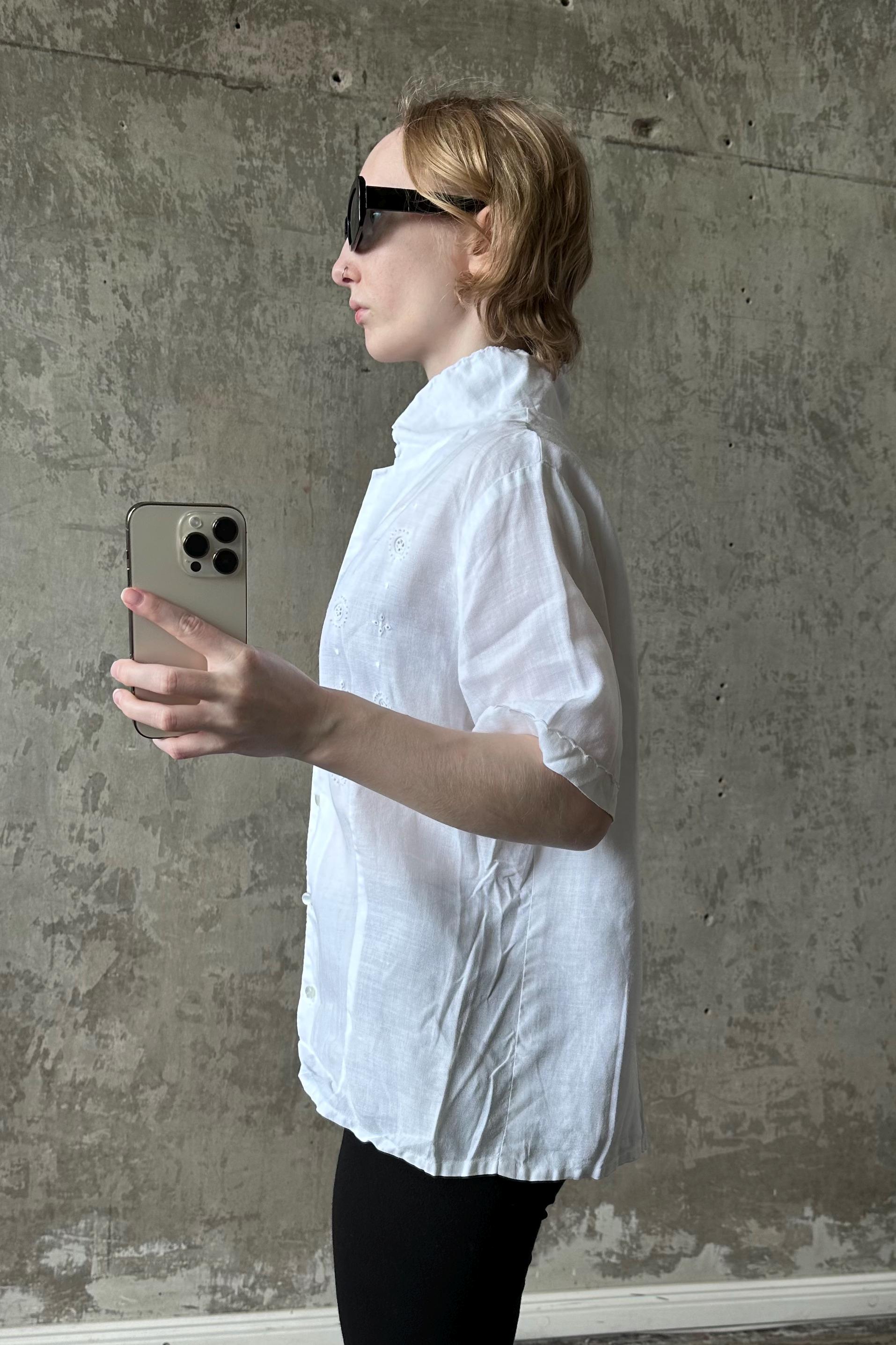 A person wearing a white shirt and sunglasses is holding a smartphone and appears to be taking a selfie in front of a concrete wall.