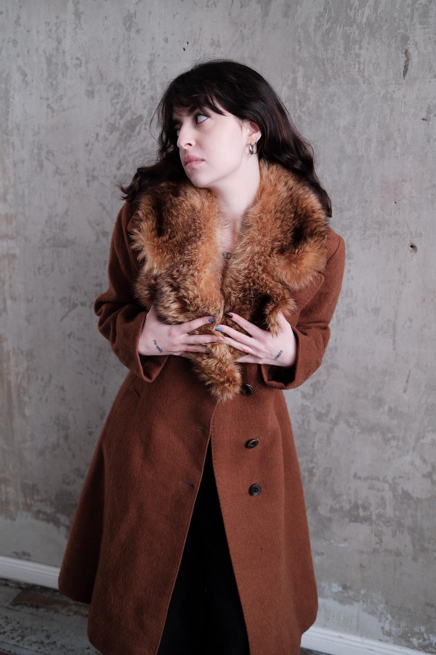A woman wearing a brown coat and fur collar is lying on the floor, looking up with a thoughtful expression.