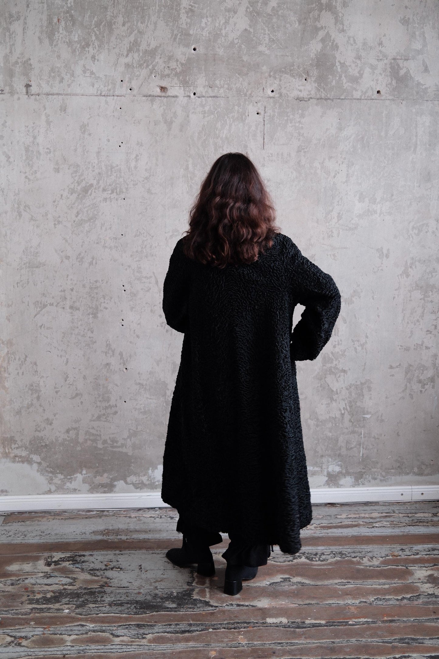 A person wearing a long black coat stands in a room with a concrete wall and wooden floor.