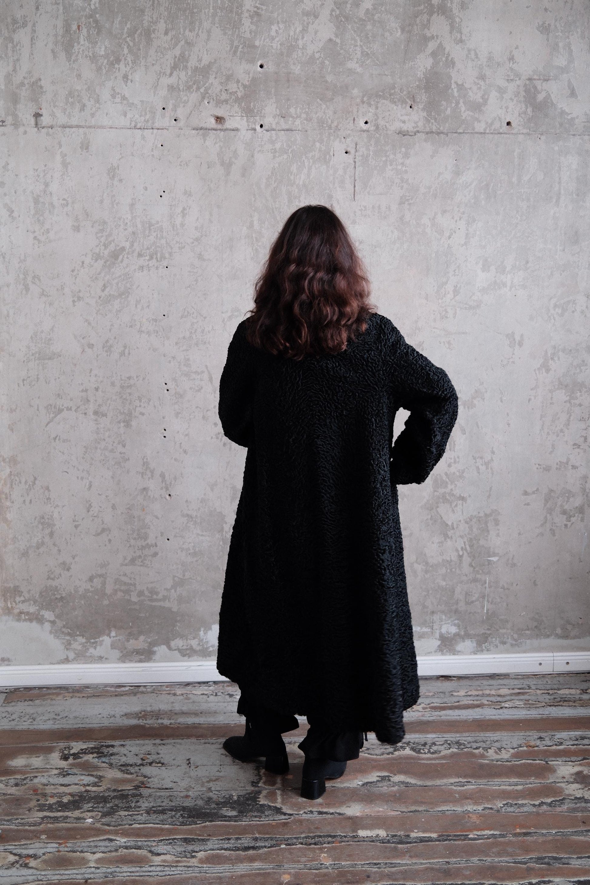 A person wearing a long black coat stands in a room with a concrete wall and wooden floor.
