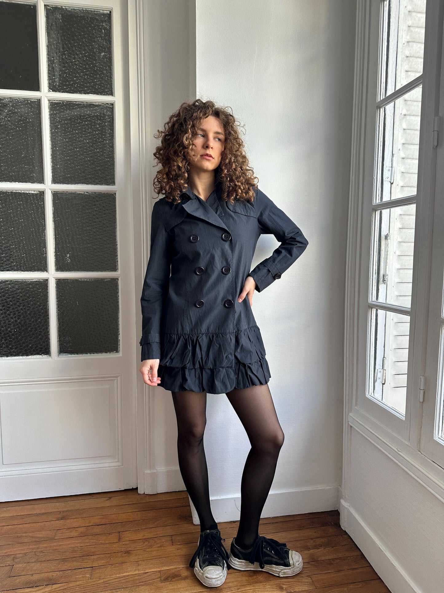 A woman with curly hair wearing a black coat and black shoes is posing in a room with a window.
