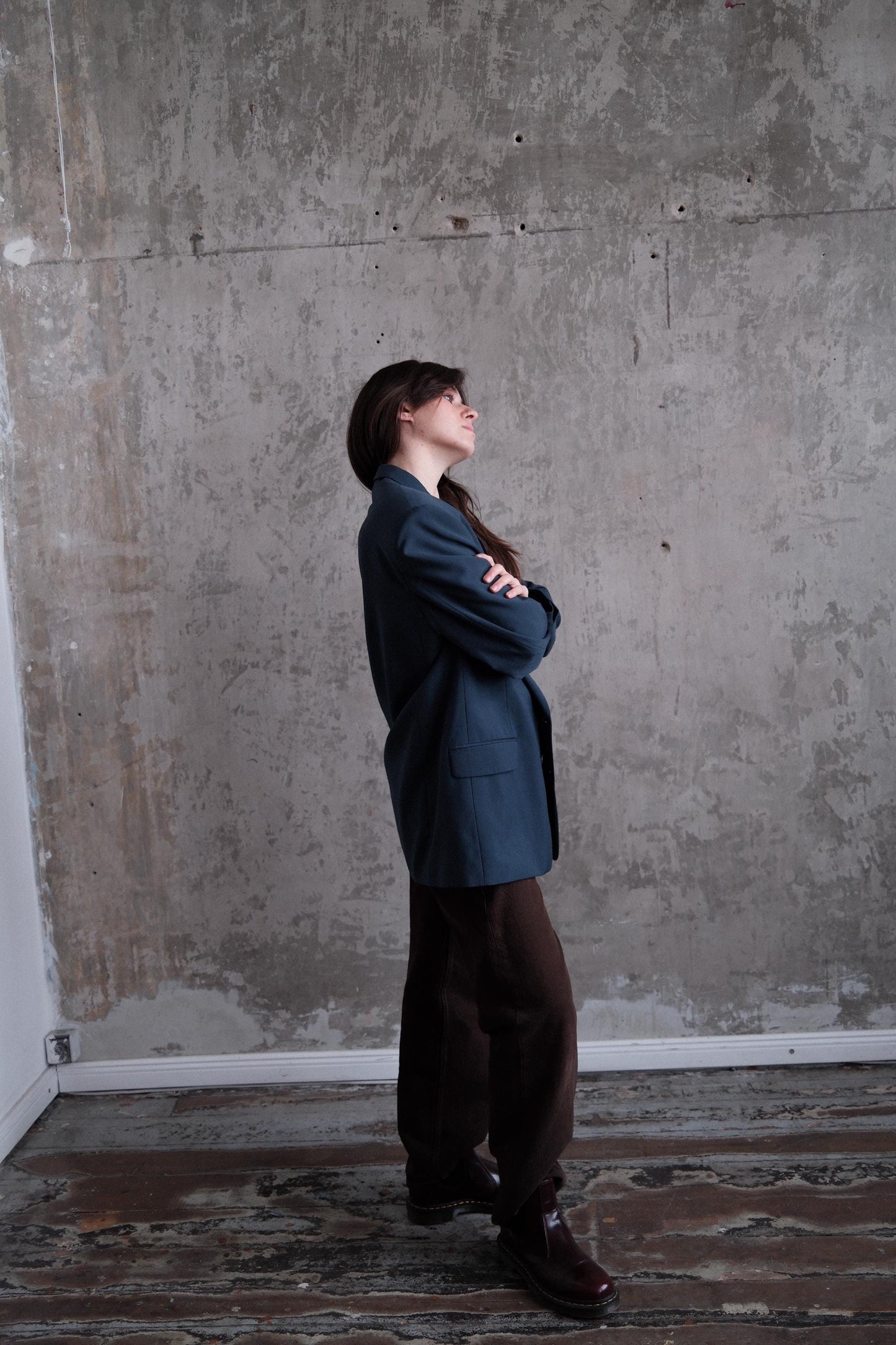 A person in a blue jacket stands with their arms crossed, looking up against a concrete wall.