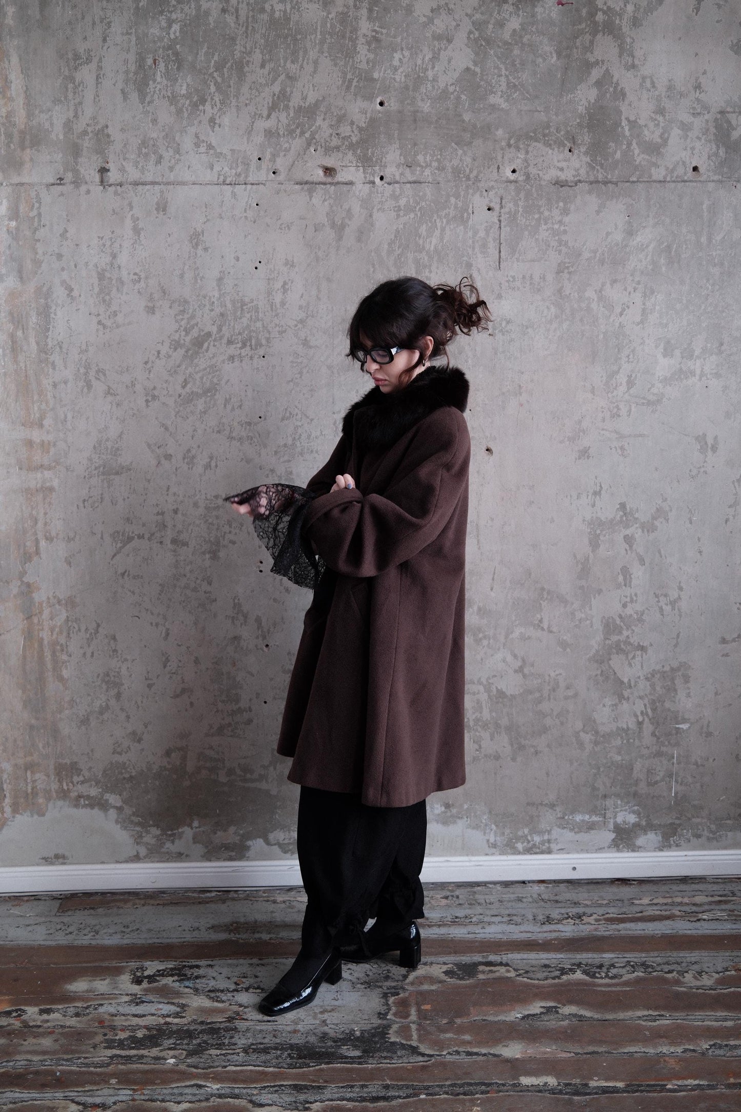 A person wearing a brown coat is standing in a room with a concrete wall.