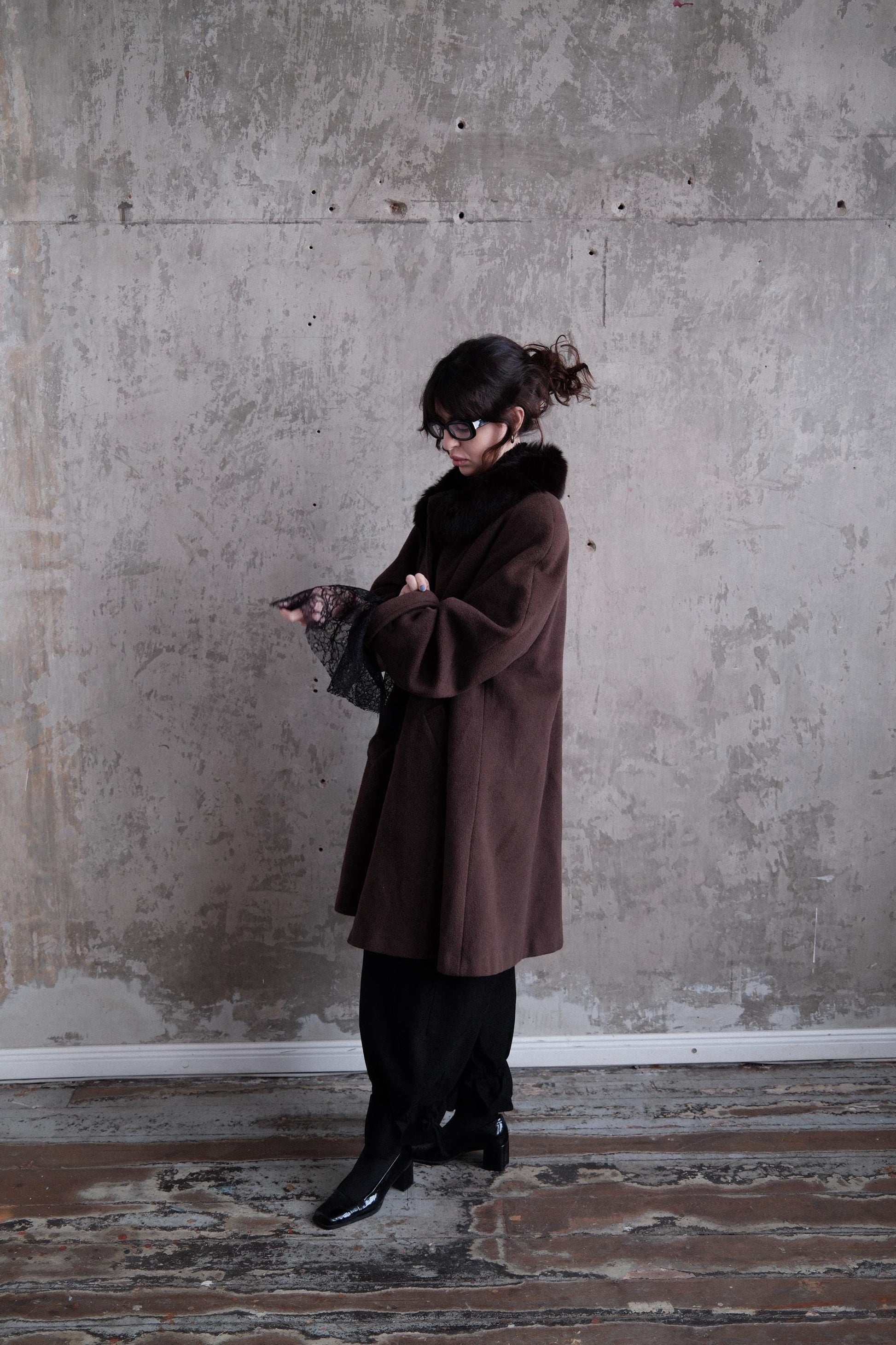 A person wearing a brown coat is standing in a room with a concrete wall.