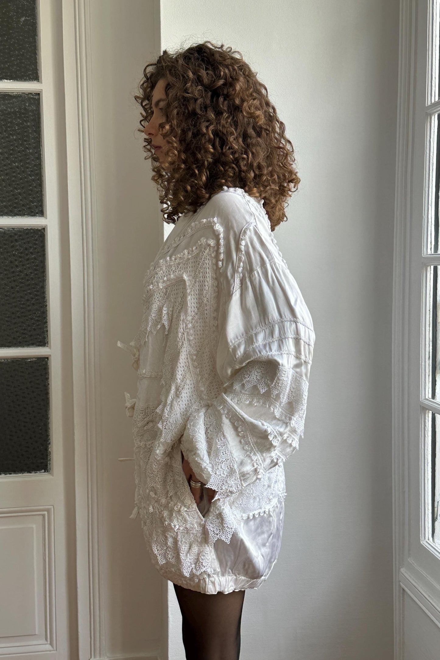 A woman with curly hair wearing a white, lace-trimmed dress stands in a room with white walls and a window.