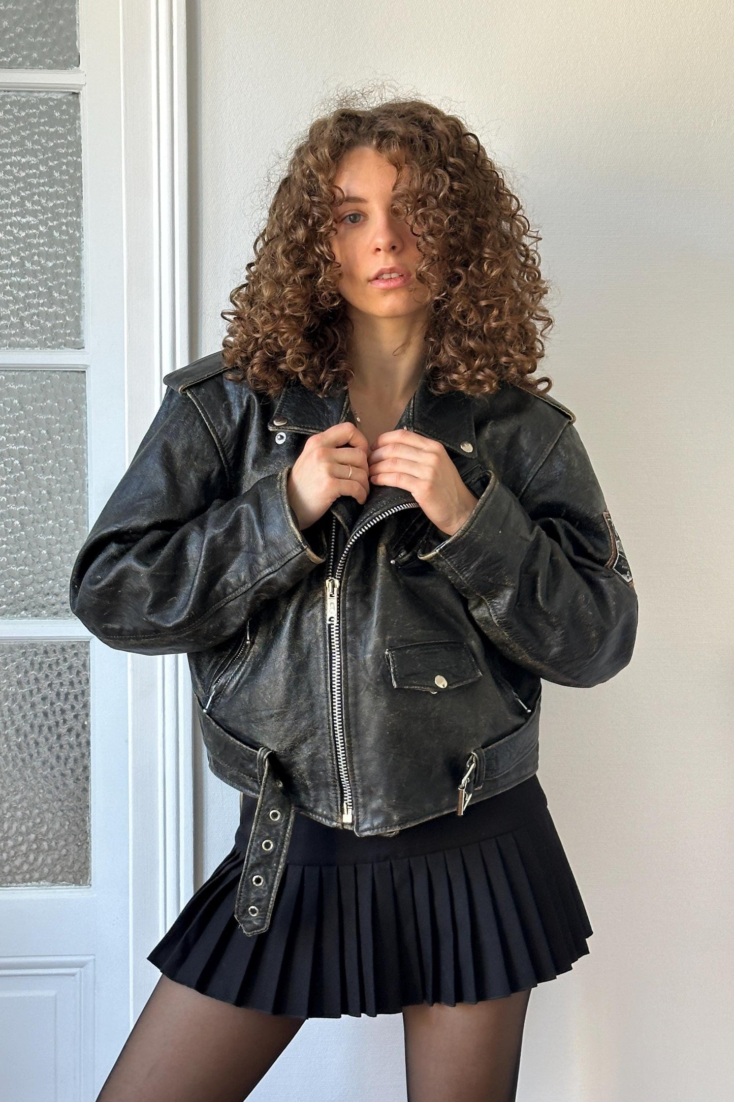 A woman with curly hair wearing a black leather jacket and a black pleated skirt stands in front of a white door.