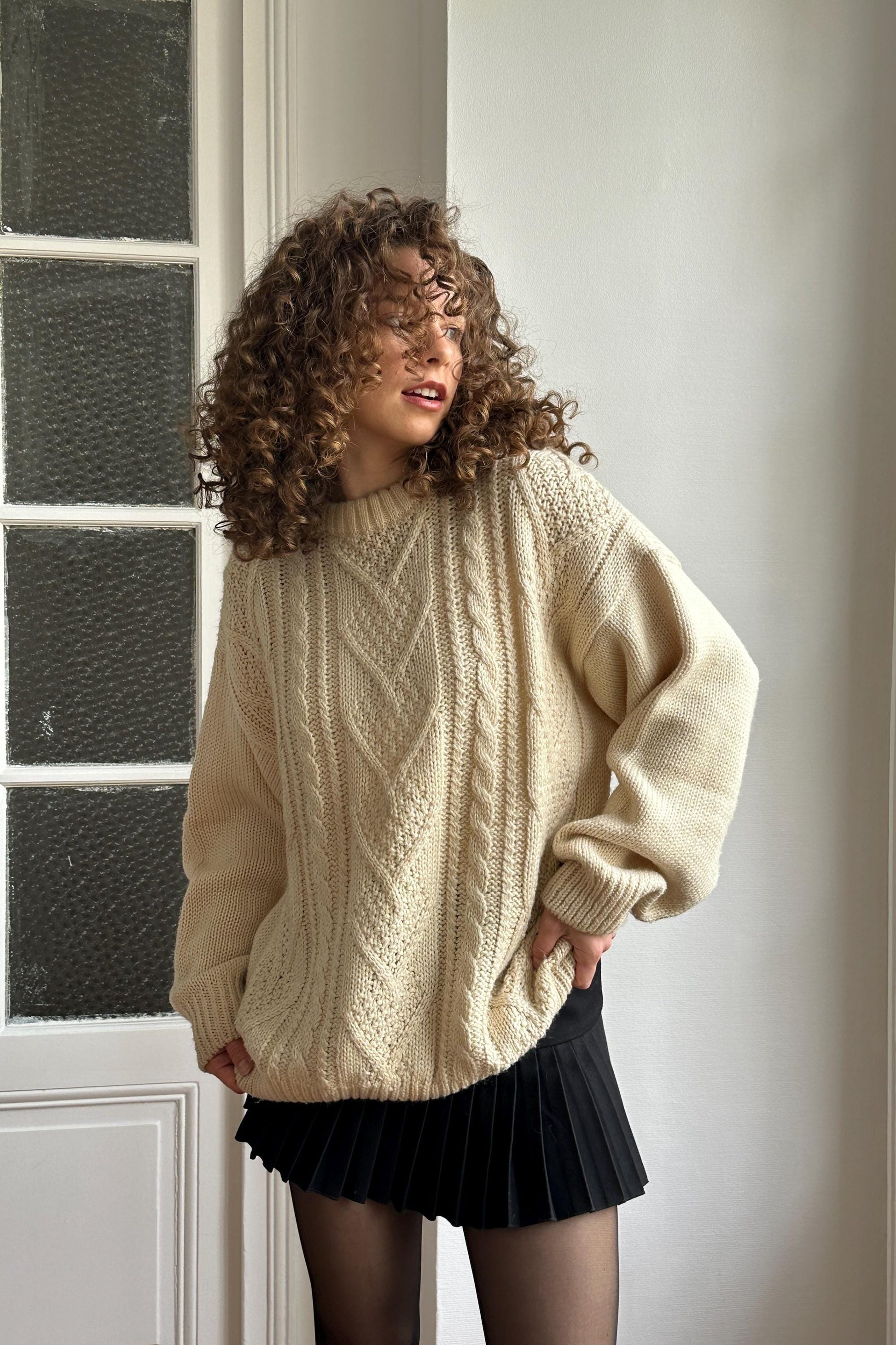 A woman with curly hair wearing a cream-colored sweater and a black skirt stands in front of a window.