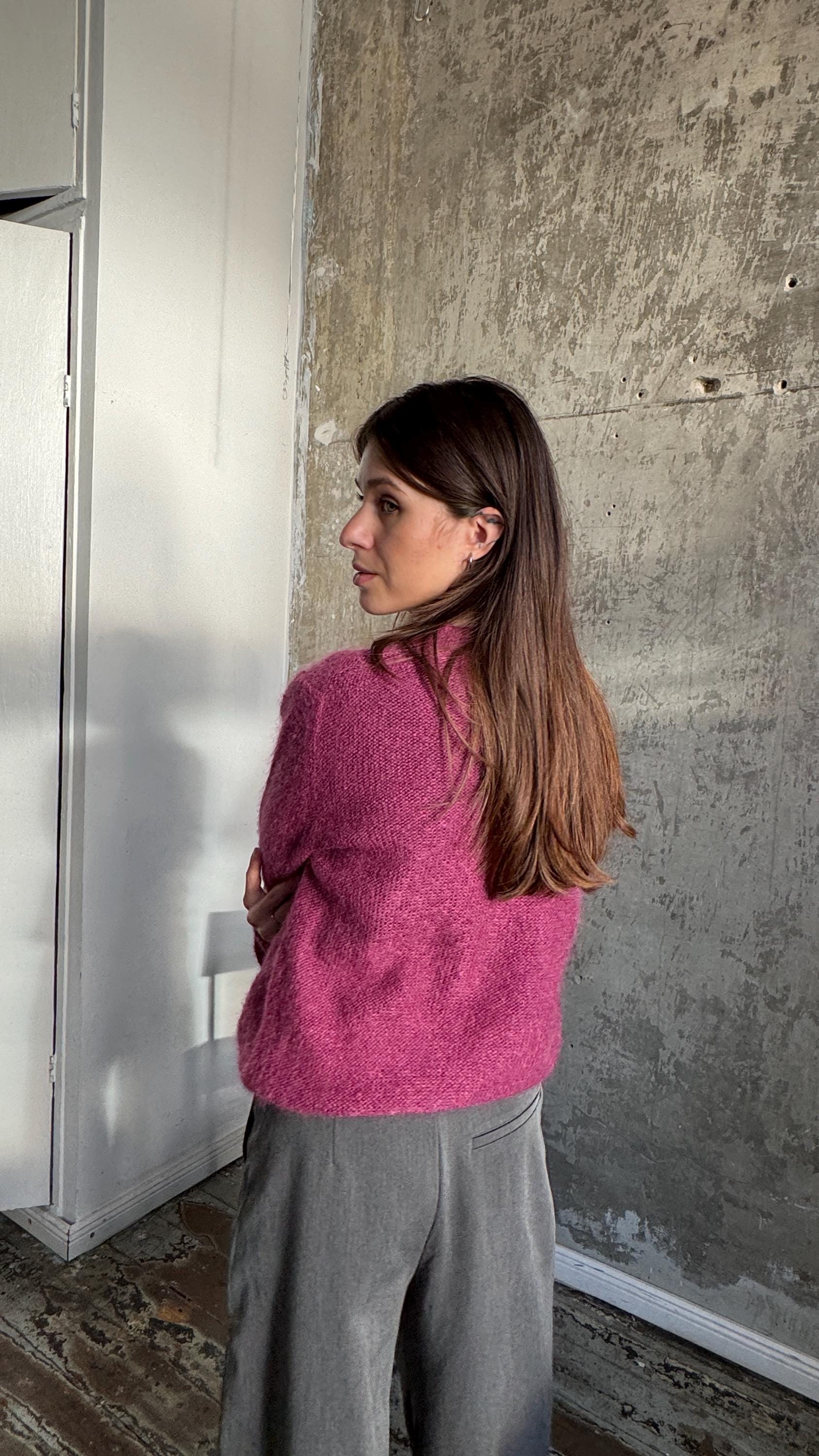 A woman with long brown hair wearing a pink sweater stands in front of a concrete wall, her back to the camera.