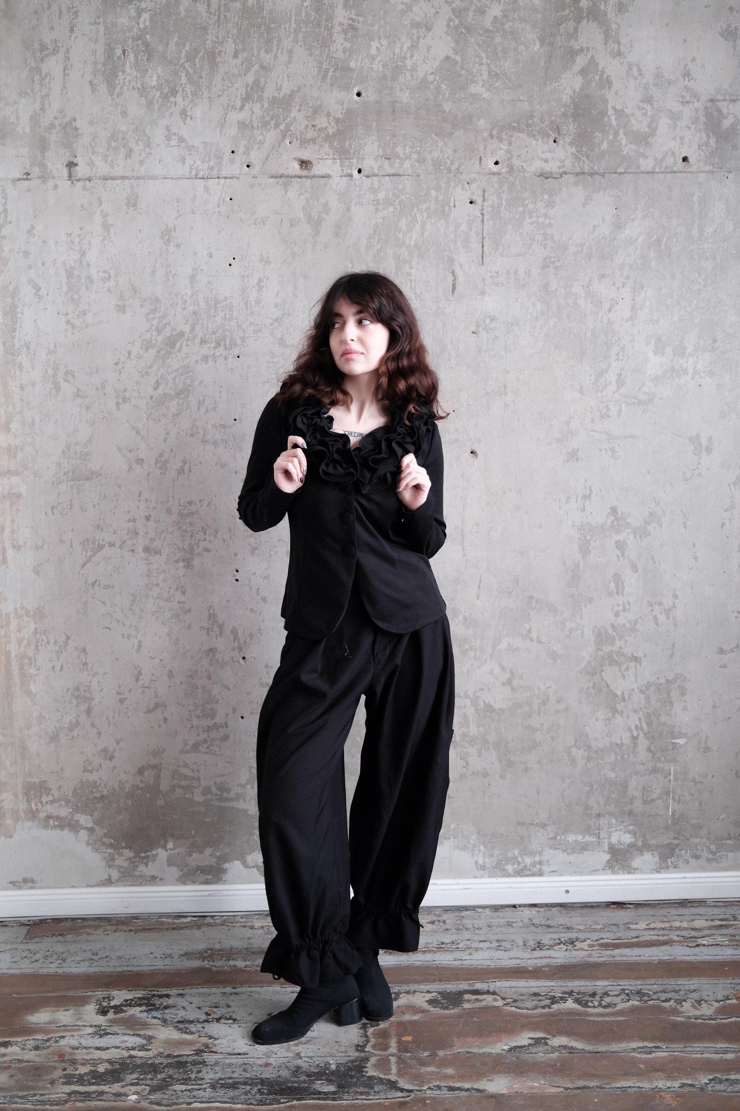 A woman in a black outfit stands in a room with a concrete wall.