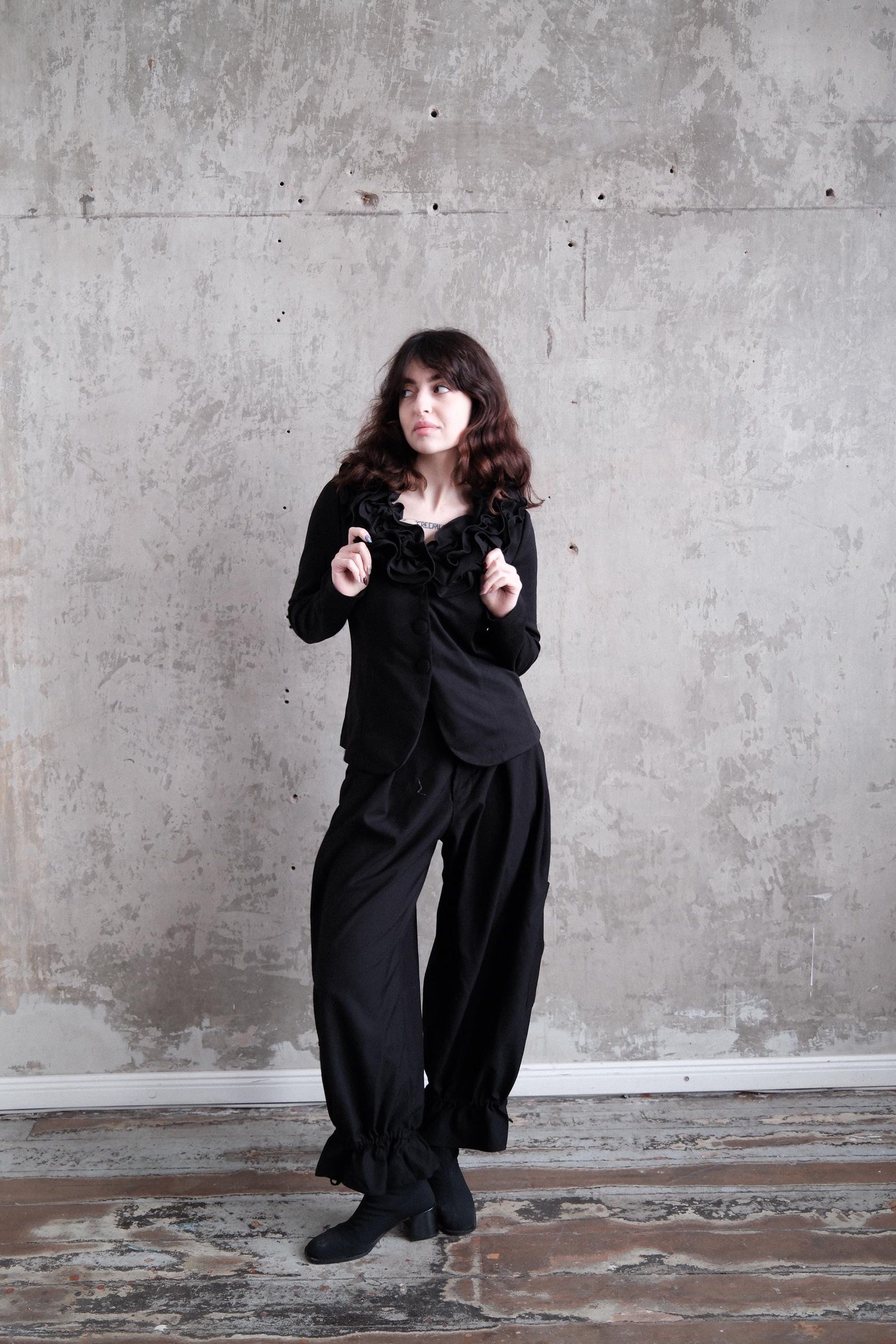 A woman in a black outfit stands in a room with a concrete wall.