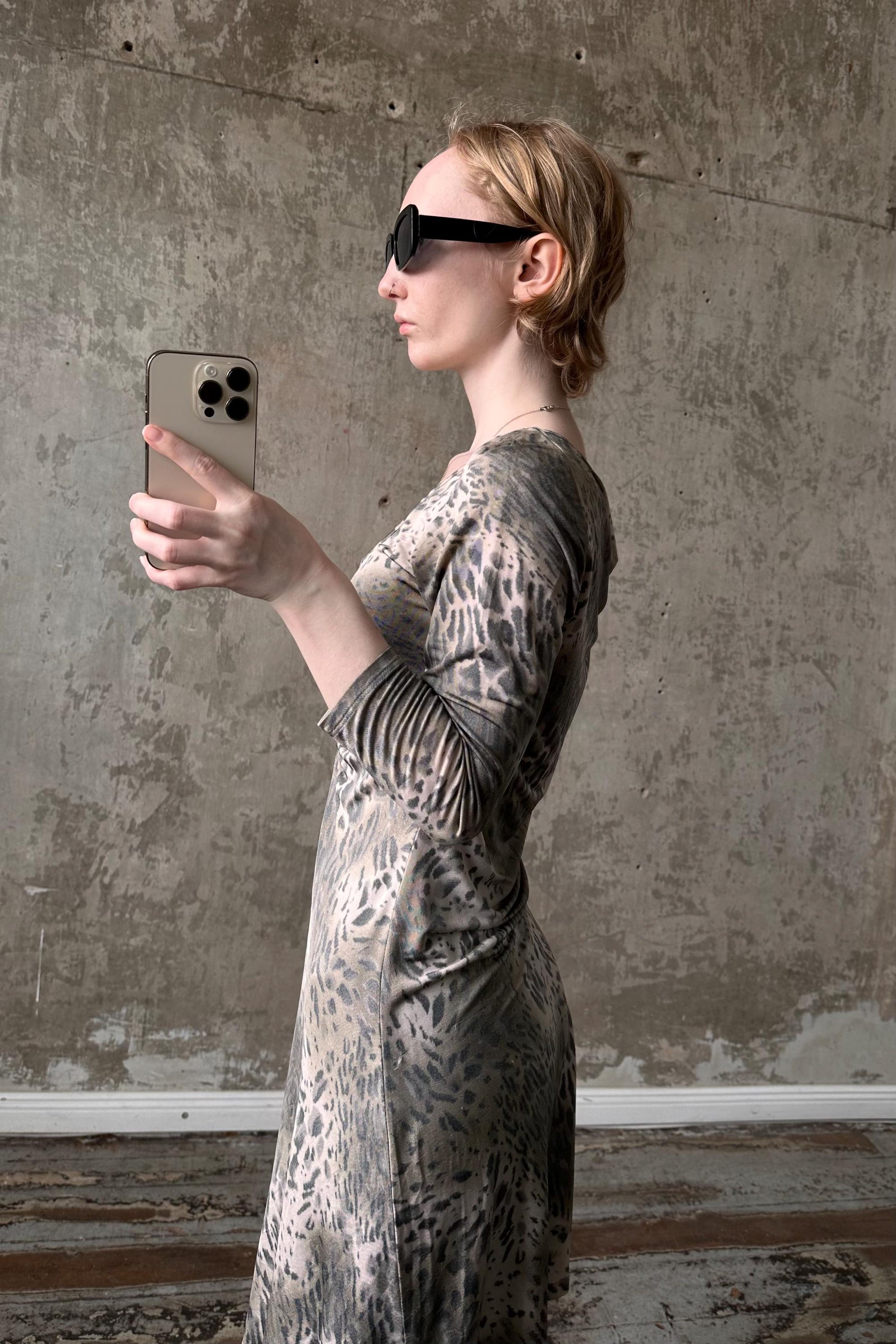 A woman wearing a leopard print dress and sunglasses is holding a smartphone and taking a selfie in front of a concrete wall.