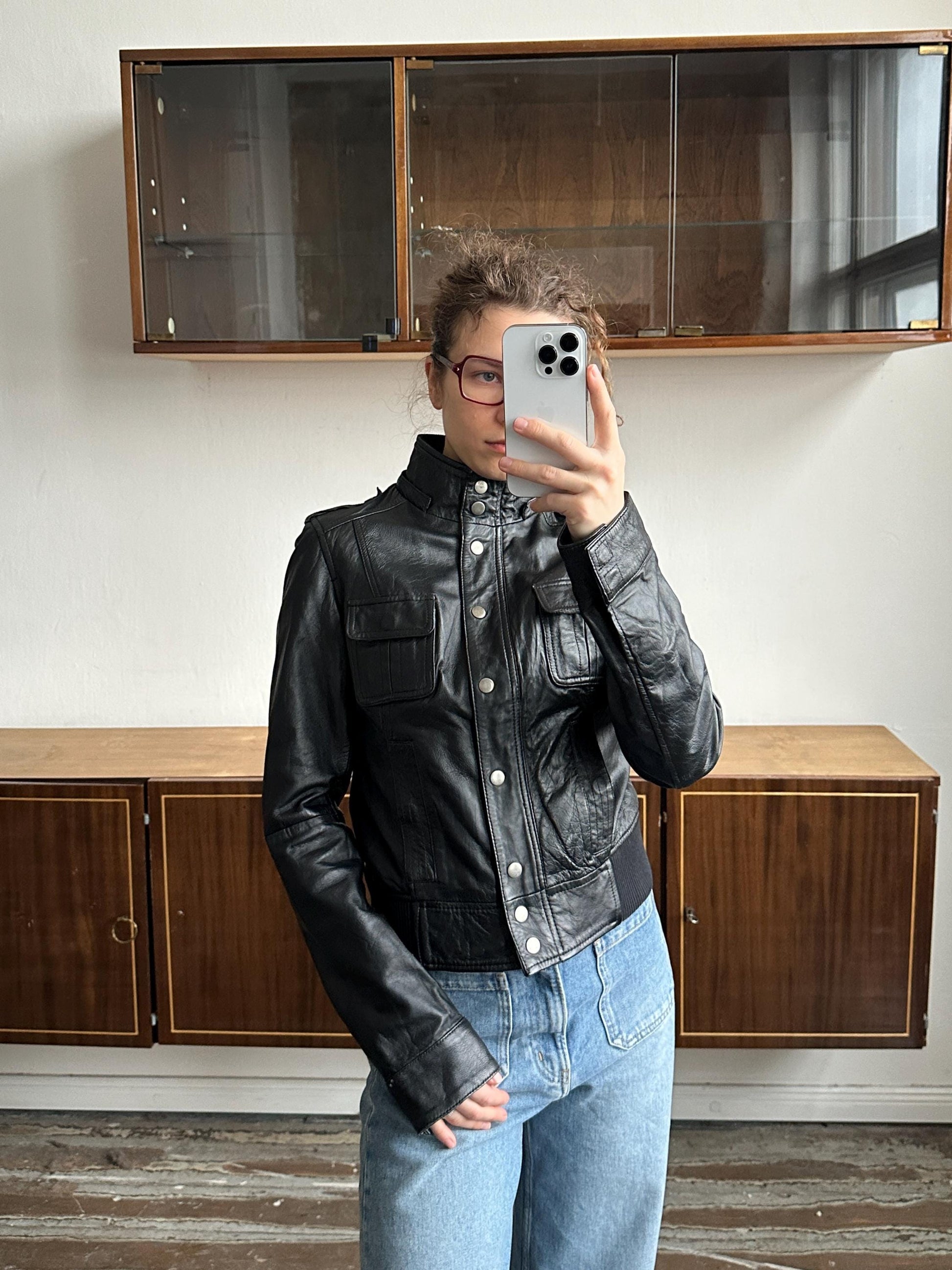 A person wearing a black leather jacket and jeans is taking a selfie with a smartphone in front of a wooden cabinet.
