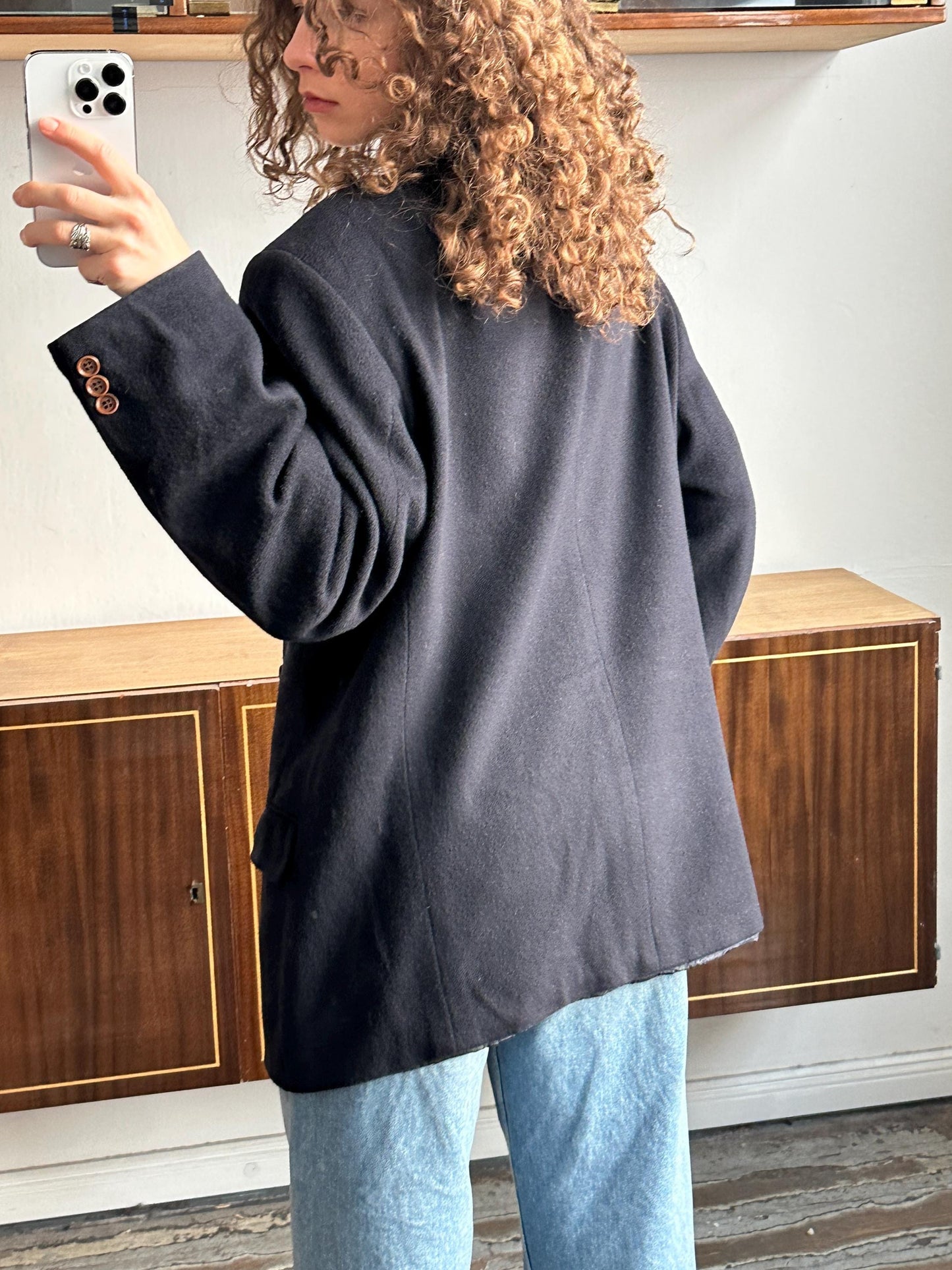 A woman with curly hair is taking a selfie with a smartphone, wearing a black blazer and blue jeans.