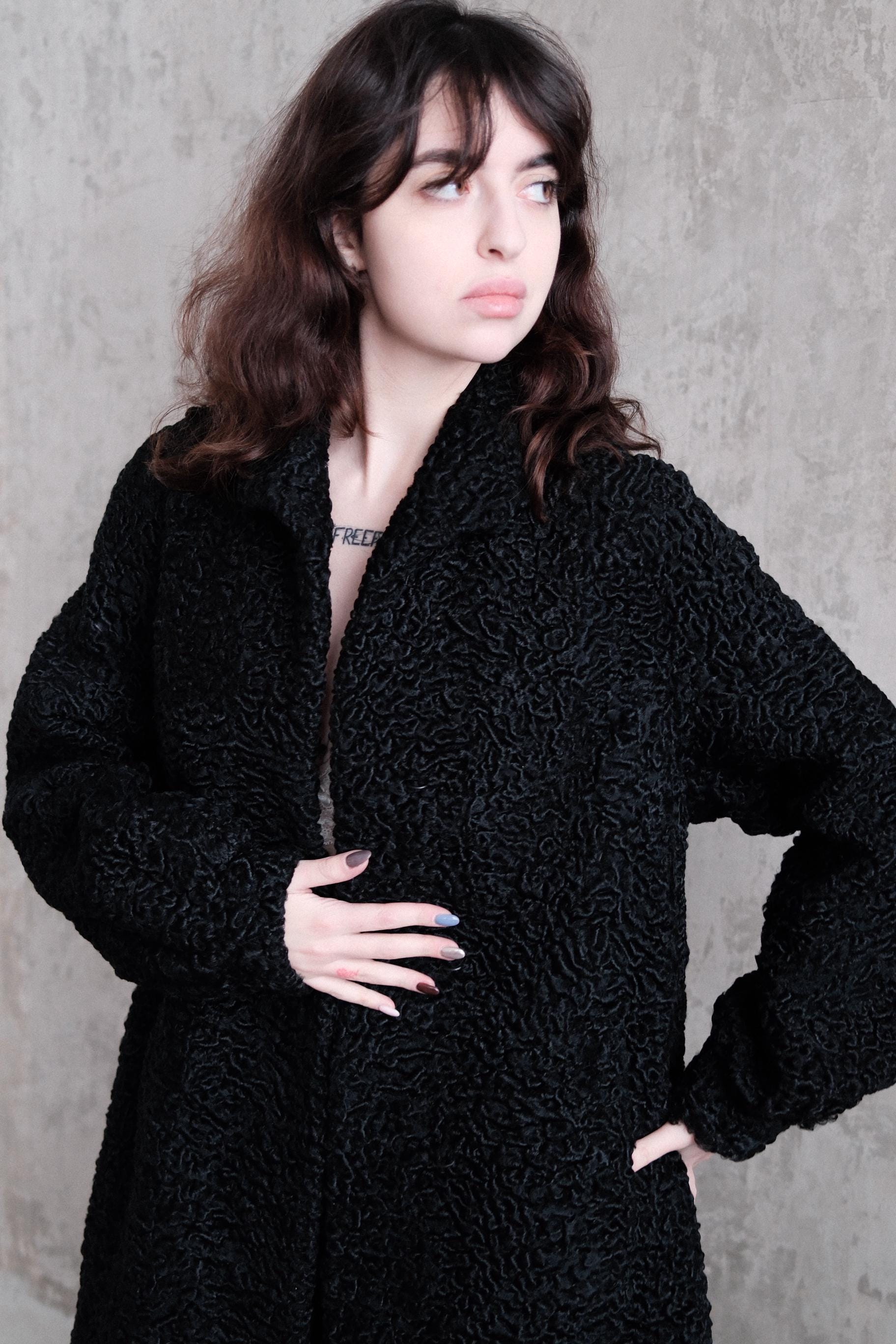 A woman wearing a black coat is posing in a relaxed manner, with her hand resting on her hip.
