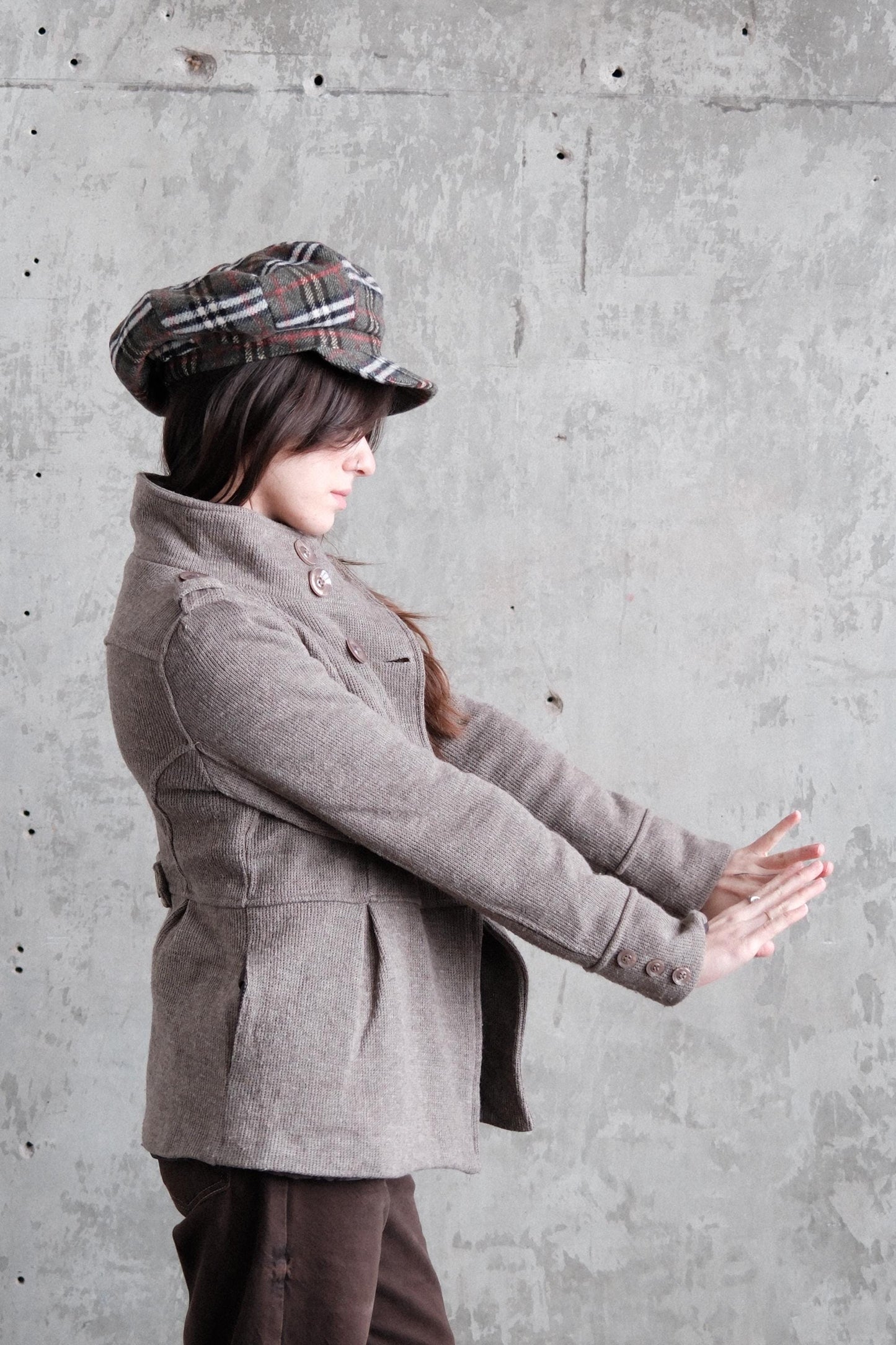 A woman in a gray coat and plaid hat is leaning over a concrete wall, with her hands clasped together.