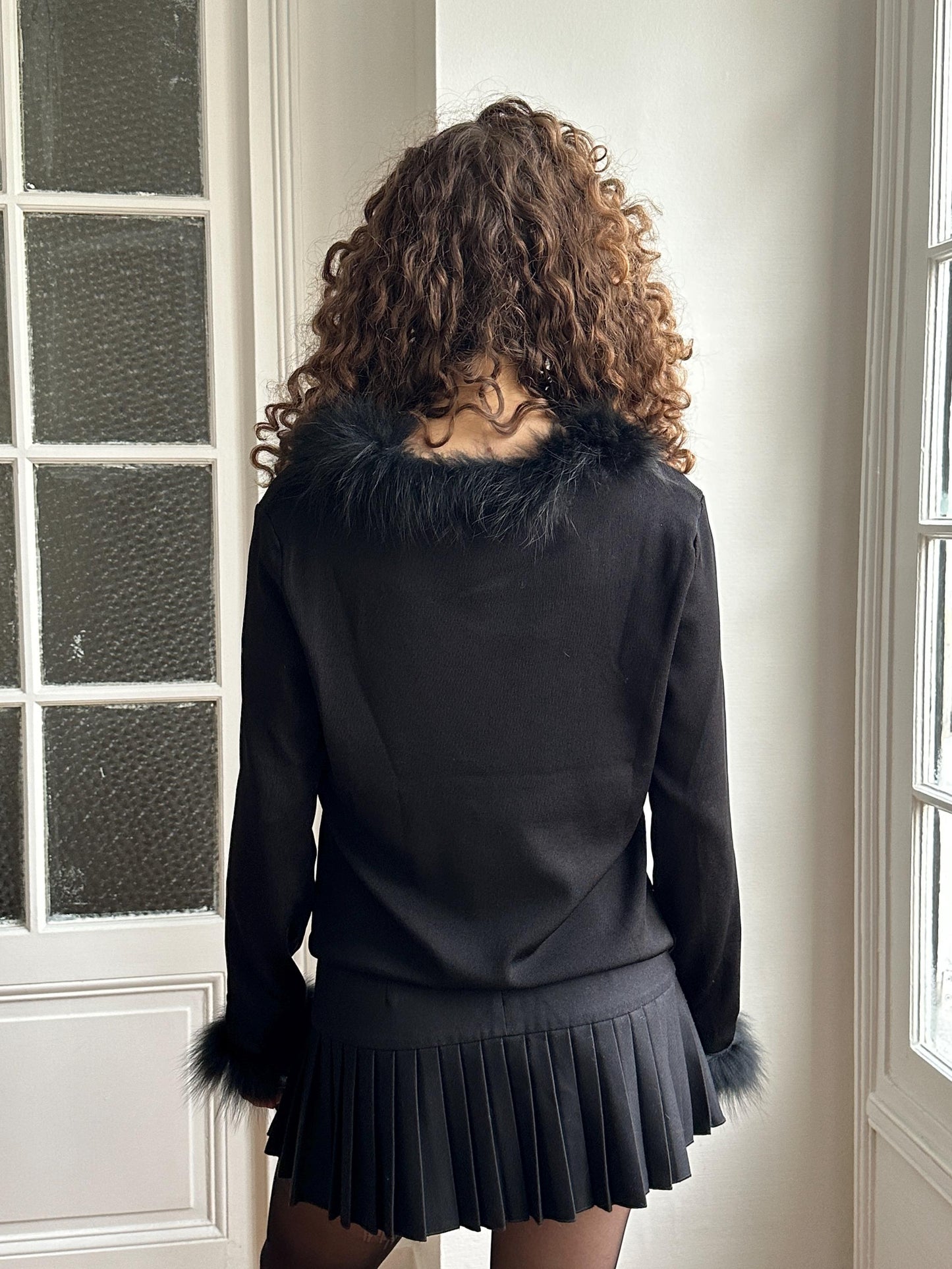 A woman with curly hair wearing a black fur collar and a black skirt stands in front of a window.