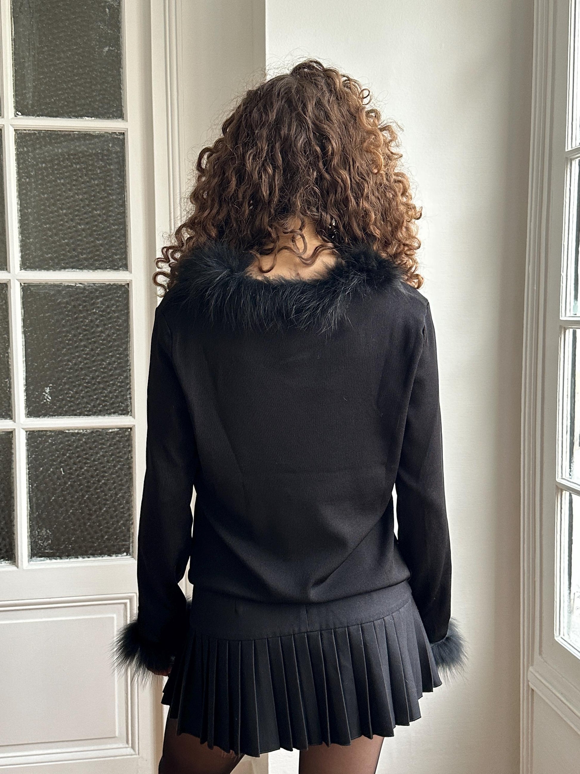 A woman with curly hair wearing a black fur collar and a black skirt stands in front of a window.