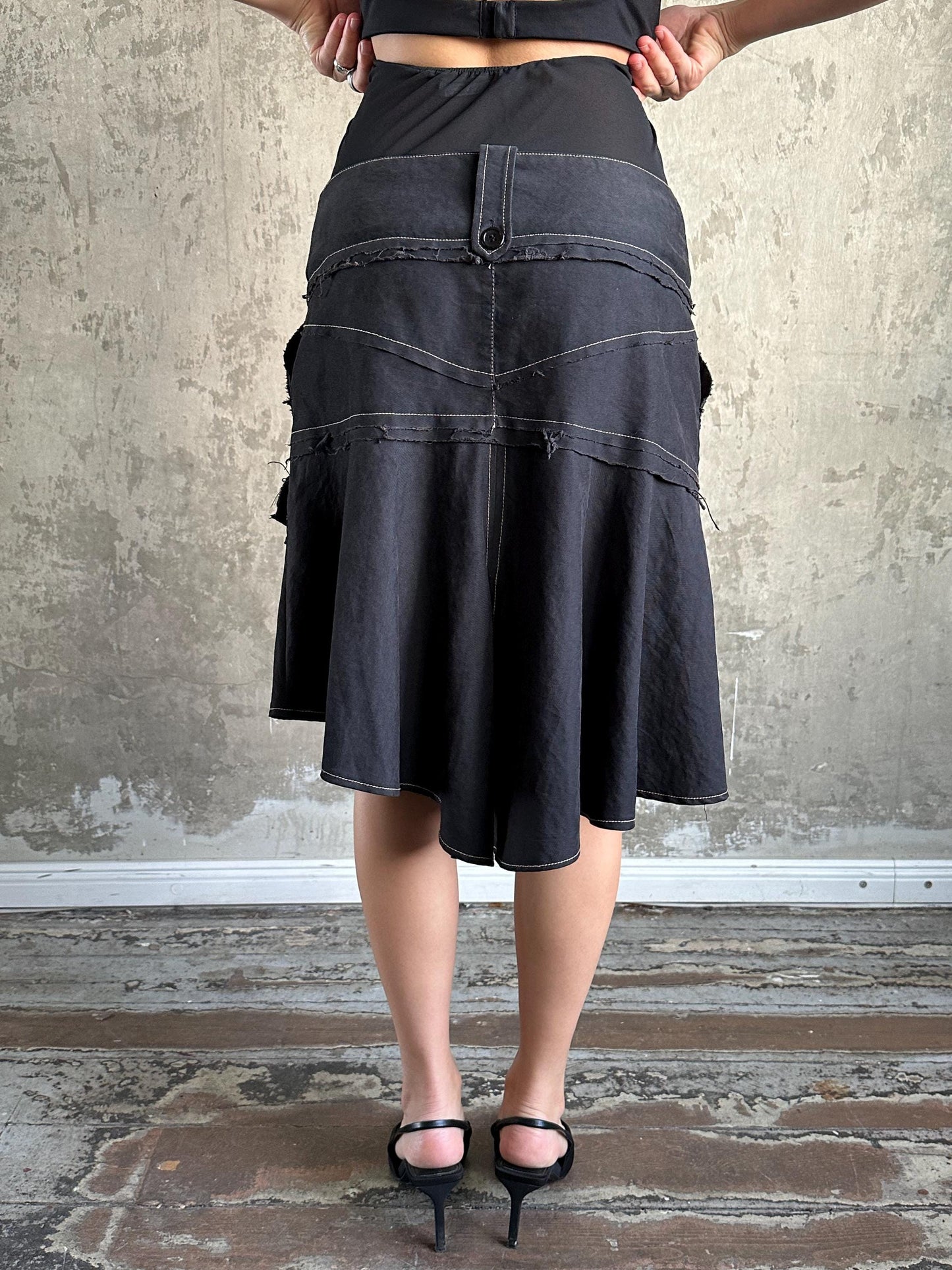 A woman wearing a black skirt with a high waist and a black top stands in front of a concrete wall.