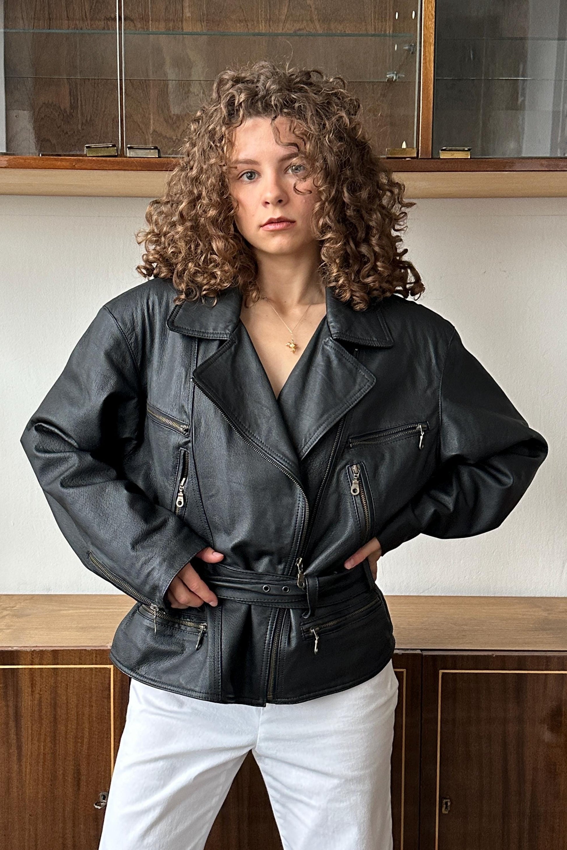 A woman with curly hair wearing a black leather jacket stands in front of a wooden cabinet.