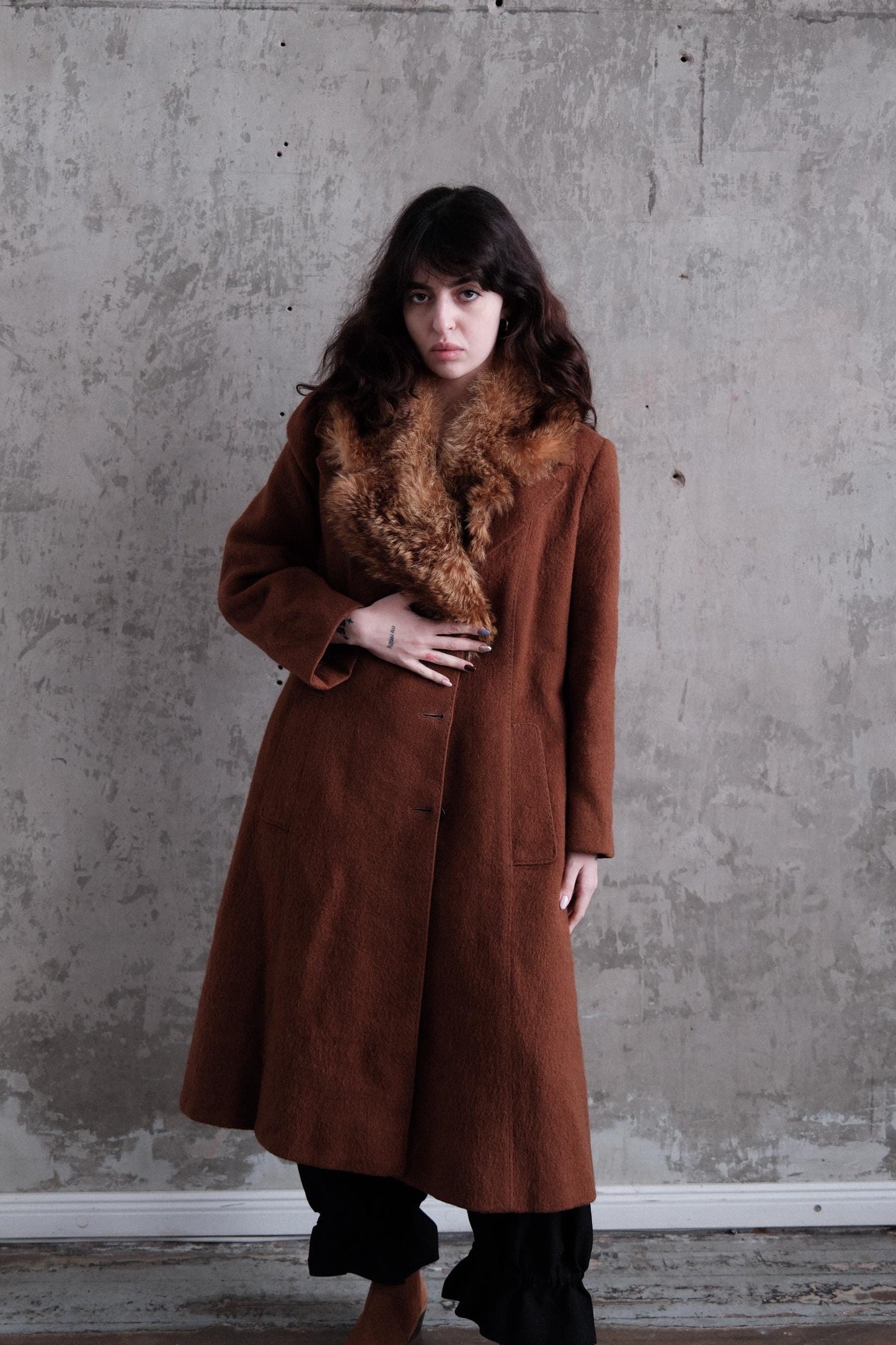 A woman wearing a brown coat with a fur collar stands against a concrete wall.