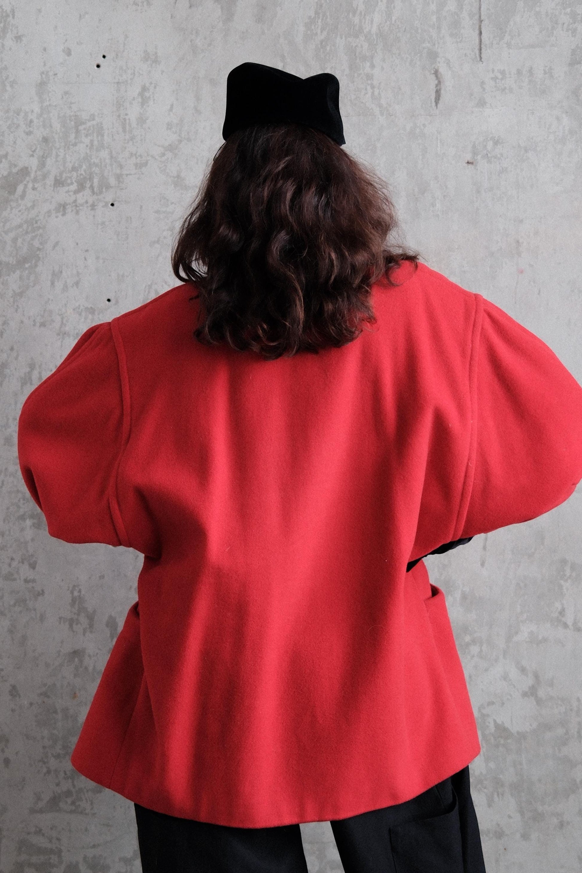 A person wearing a red jacket and black pants is standing against a concrete wall.