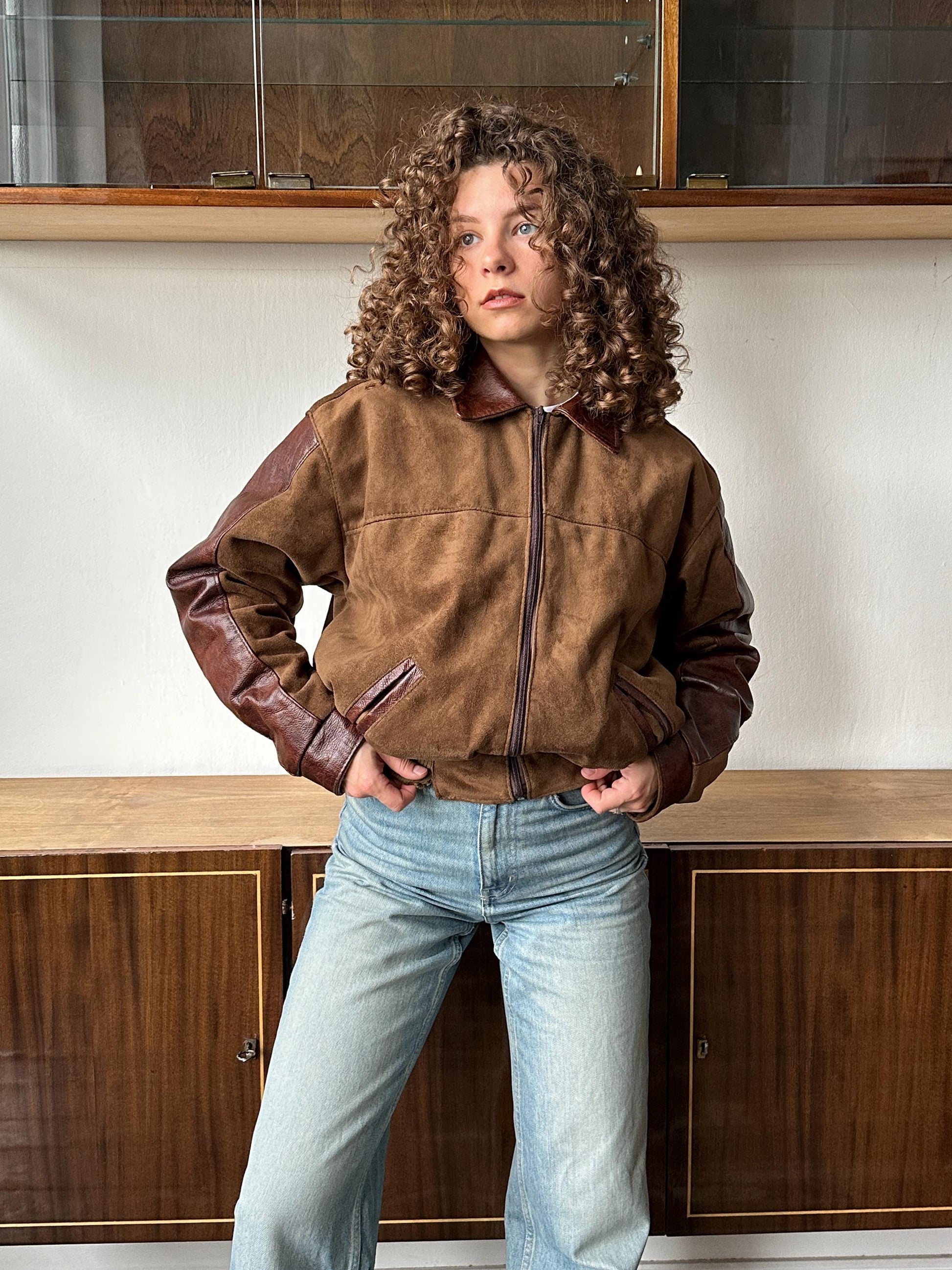 A person with curly hair wearing a brown jacket and blue jeans stands in a room with wooden cabinets in the background.