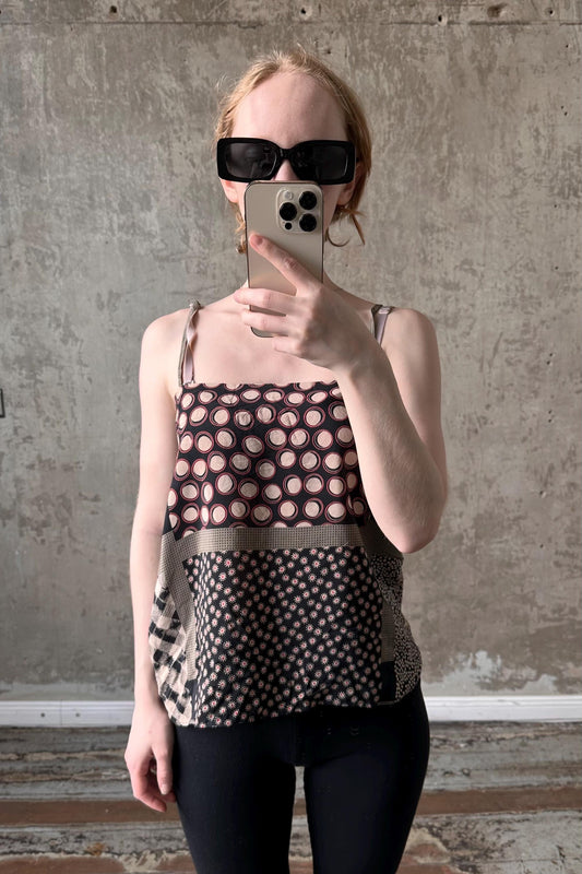 A woman wearing a patterned top and sunglasses is taking a selfie with her iPhone against a concrete wall.