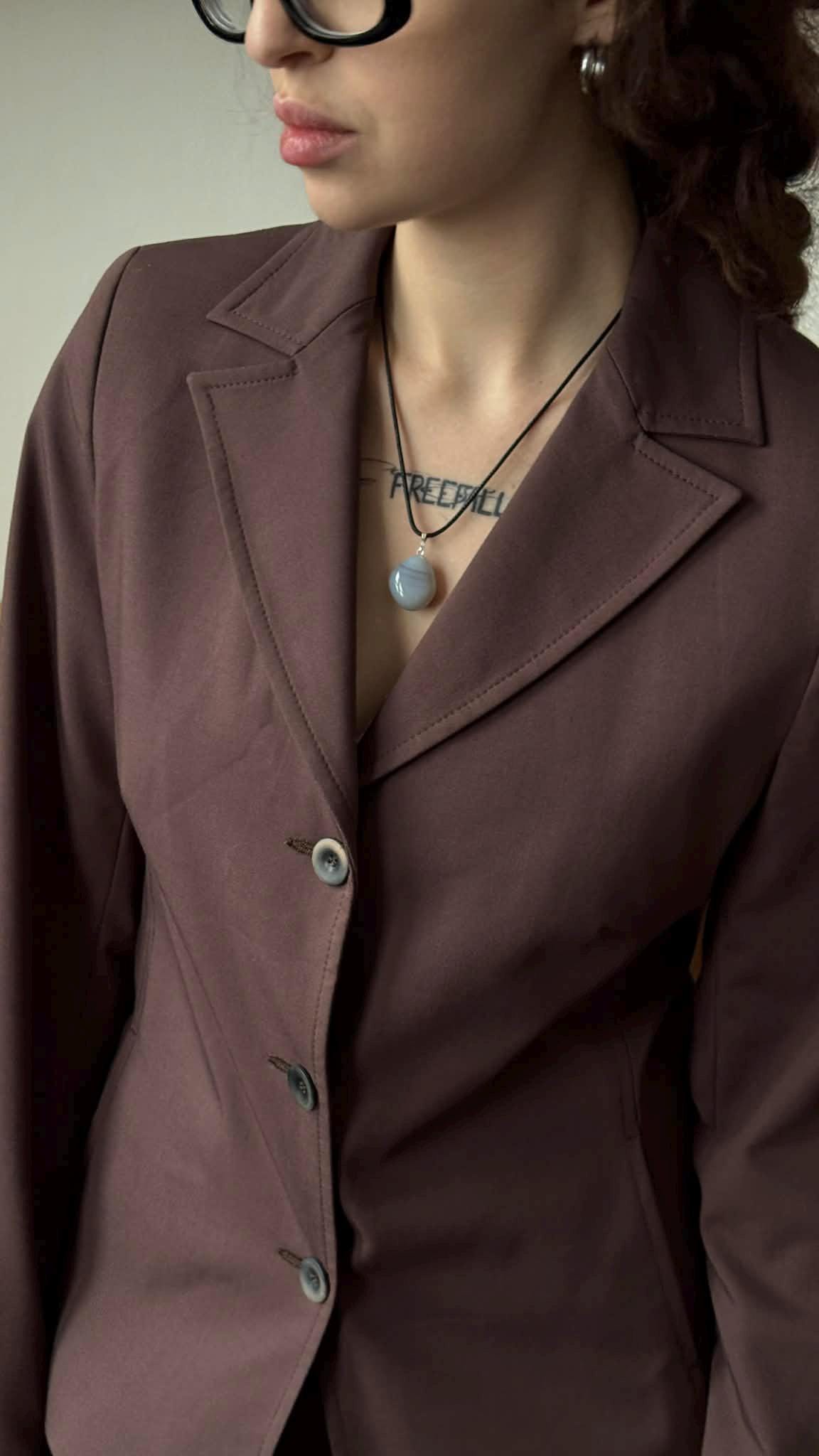 A woman wearing a brown blazer and glasses, with a necklace and a tattoo visible on her neck.