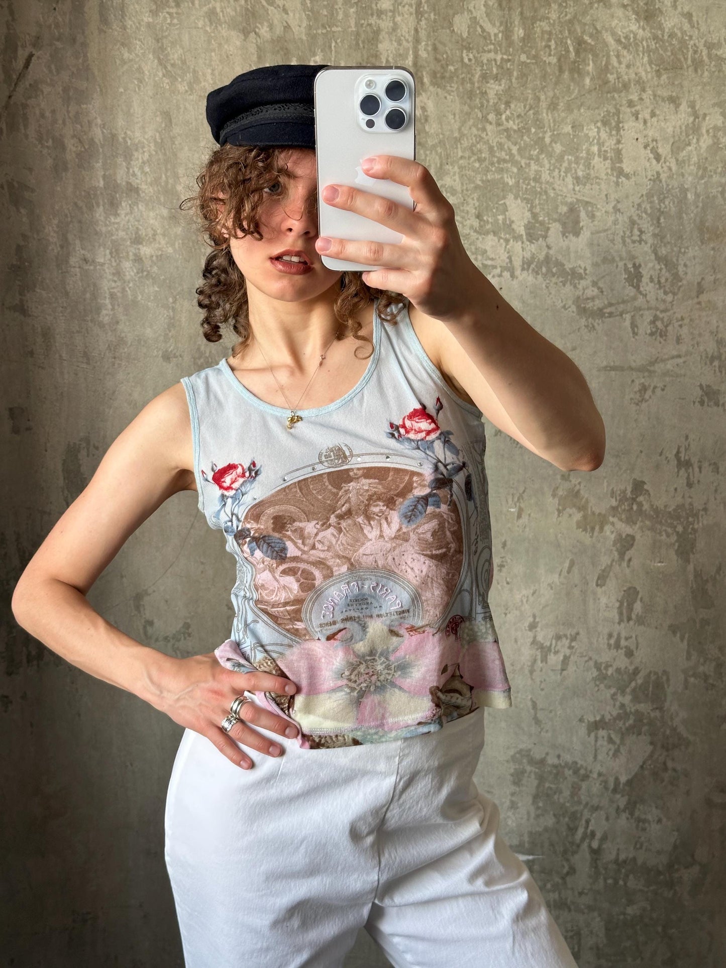 A woman is taking a selfie with a smartphone, wearing a floral tank top and white pants.