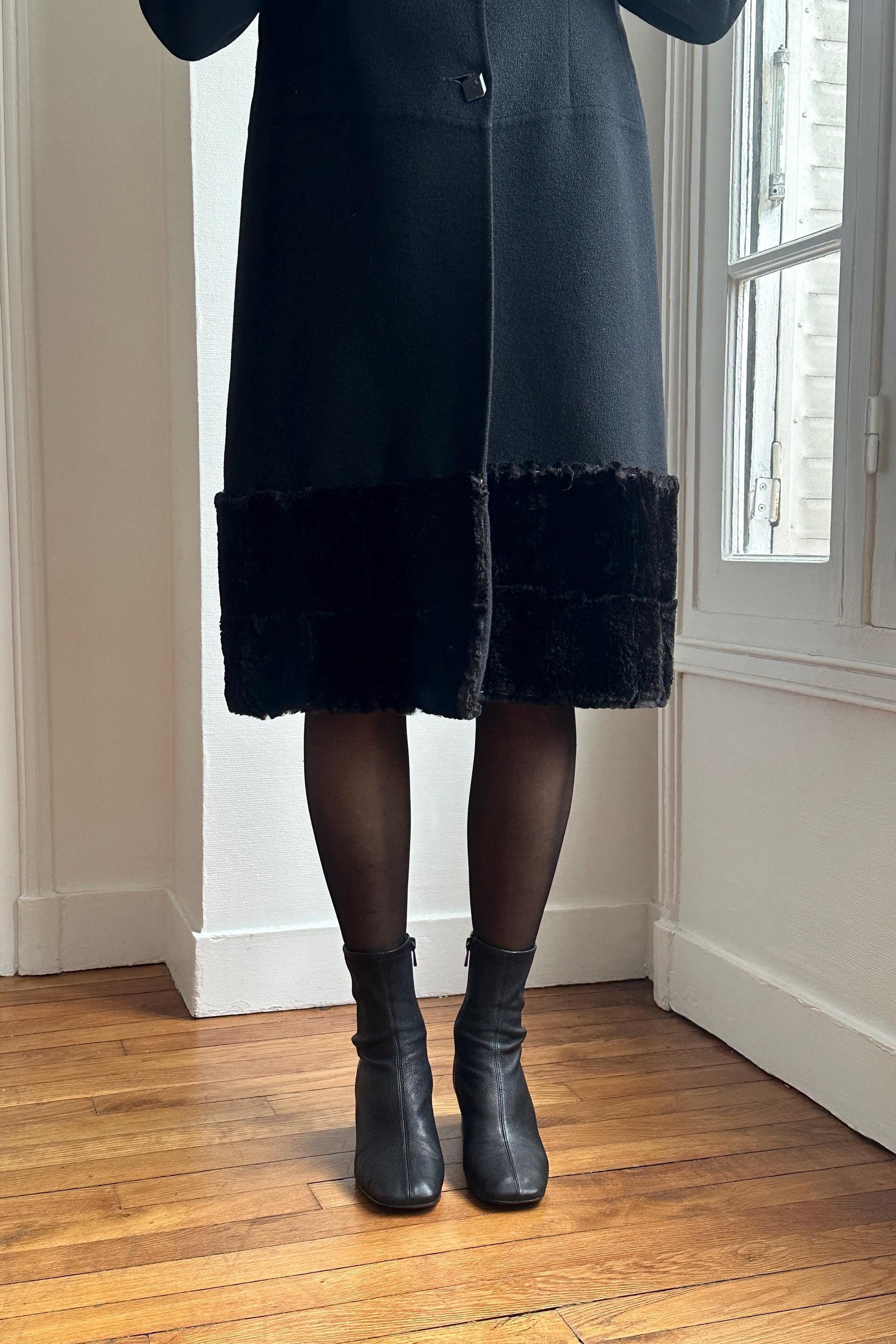 A person wearing a black coat with fur trim and black boots stands in a room with a wooden floor.