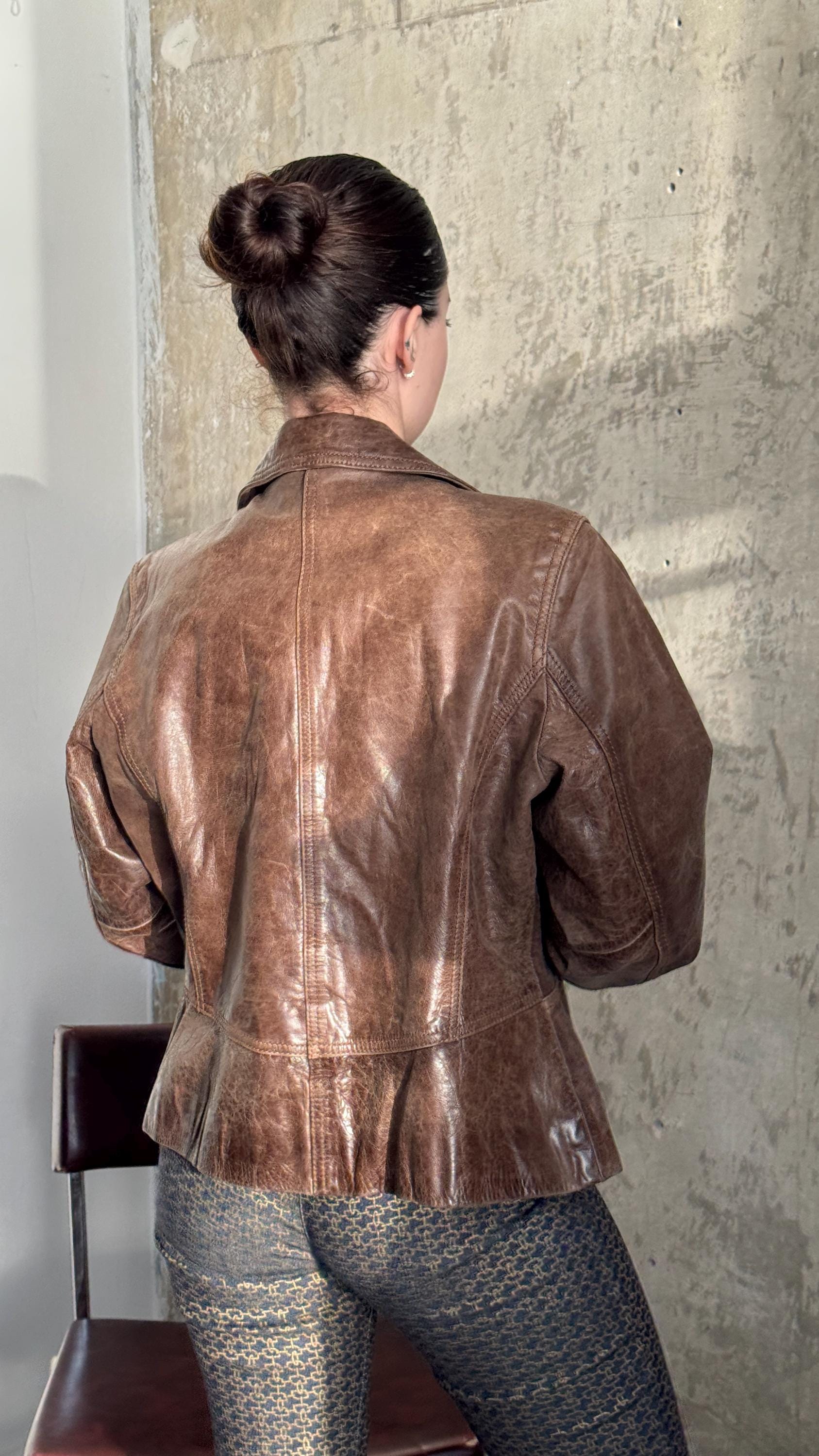 A woman wearing a brown leather jacket stands in front of a concrete wall, facing away from the camera.