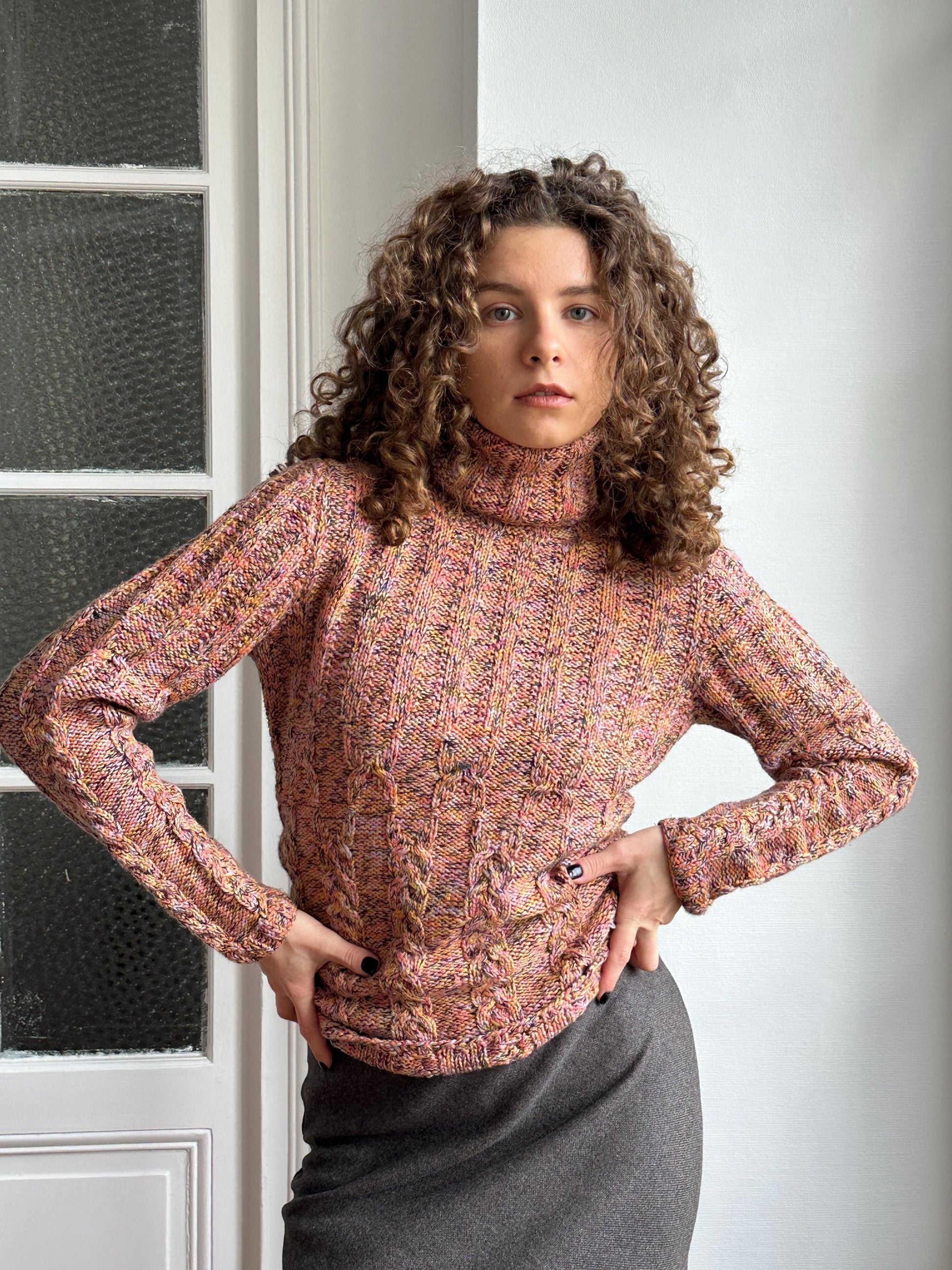 A woman with curly hair wearing a pink and orange sweater is posing with her hands on her hips, leaning against a white wall.