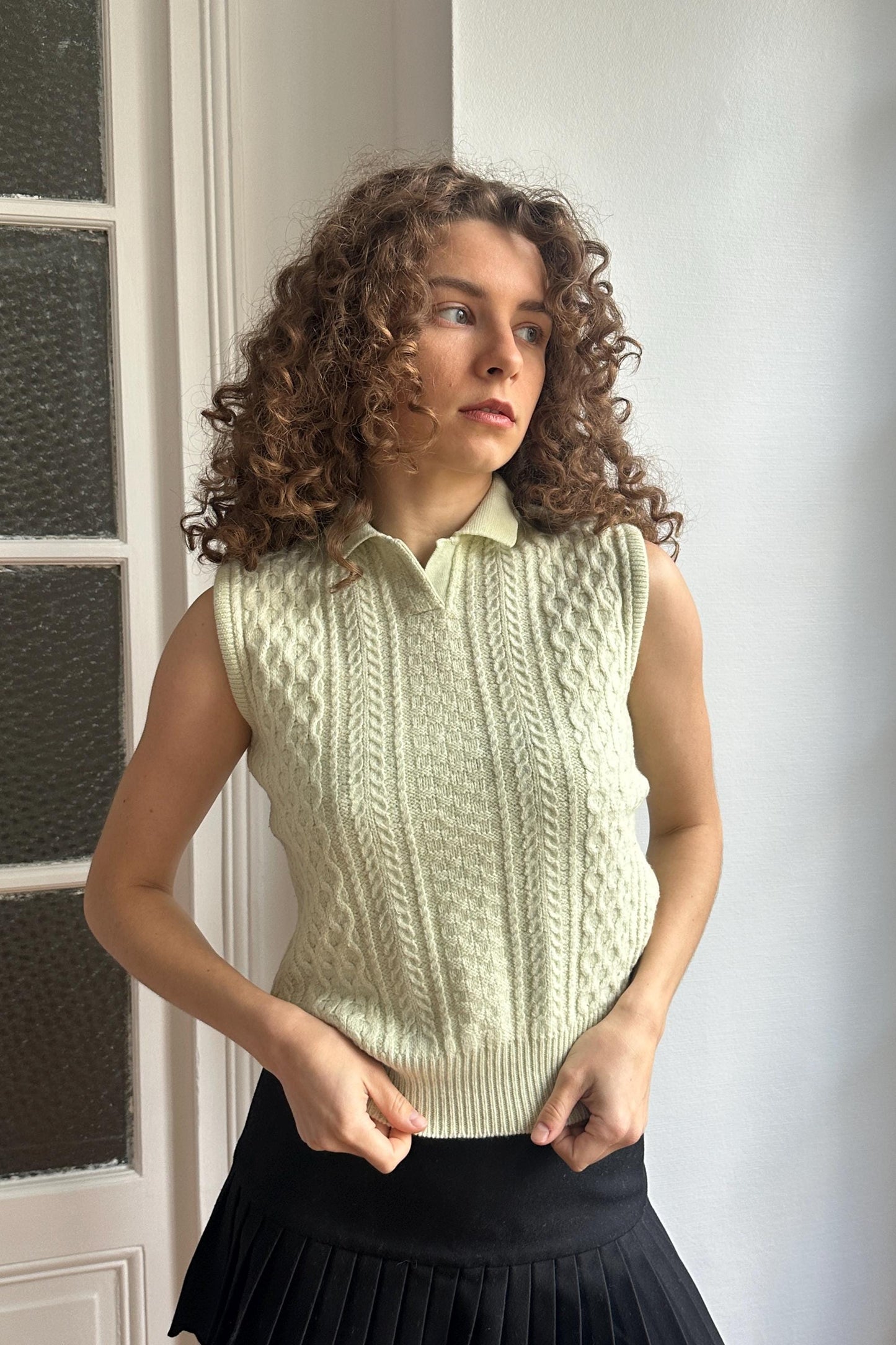 A young woman with curly hair stands in front of a white door, wearing a sleeveless white sweater and a black skirt.