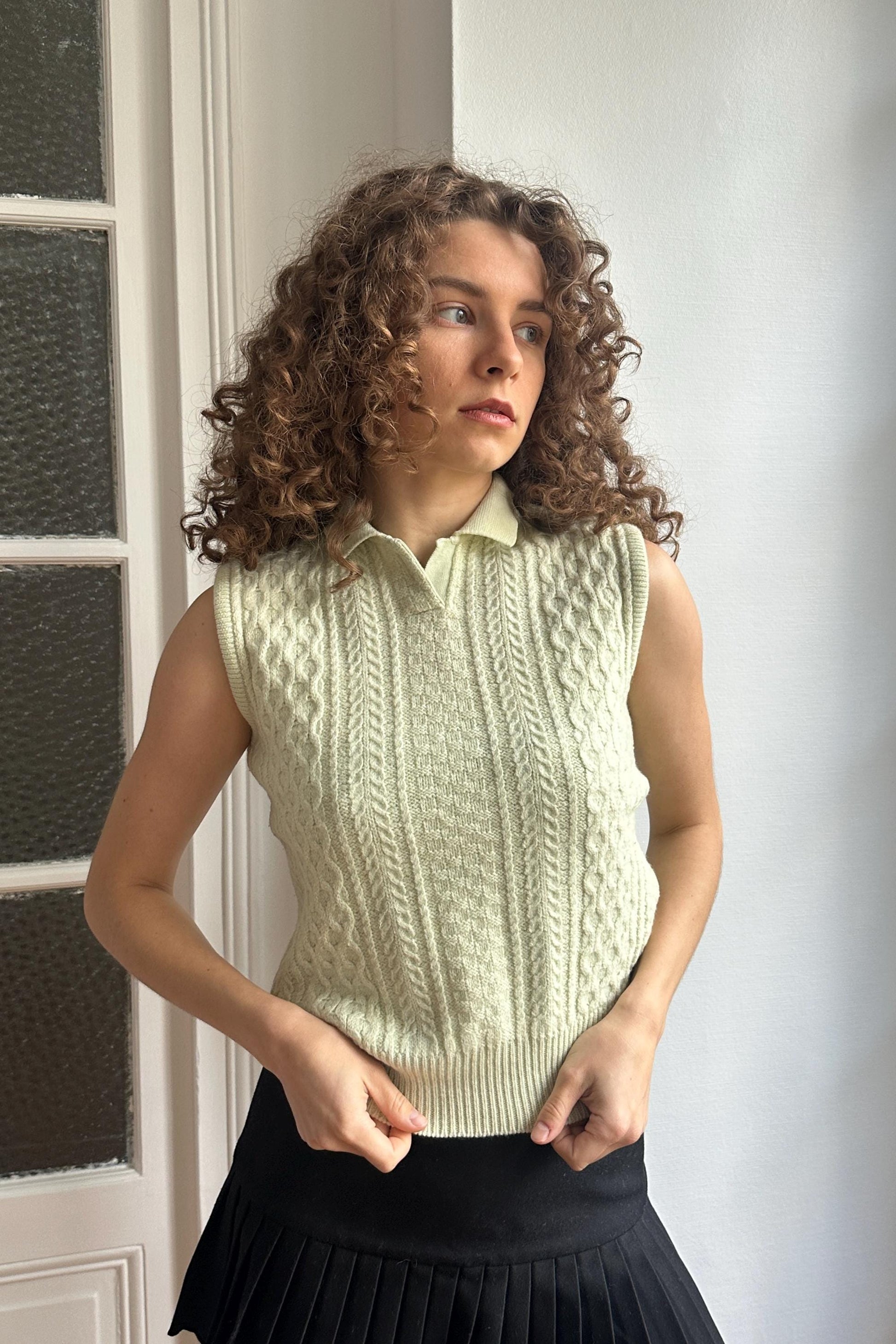A young woman with curly hair stands in front of a white door, wearing a sleeveless white sweater and a black skirt.