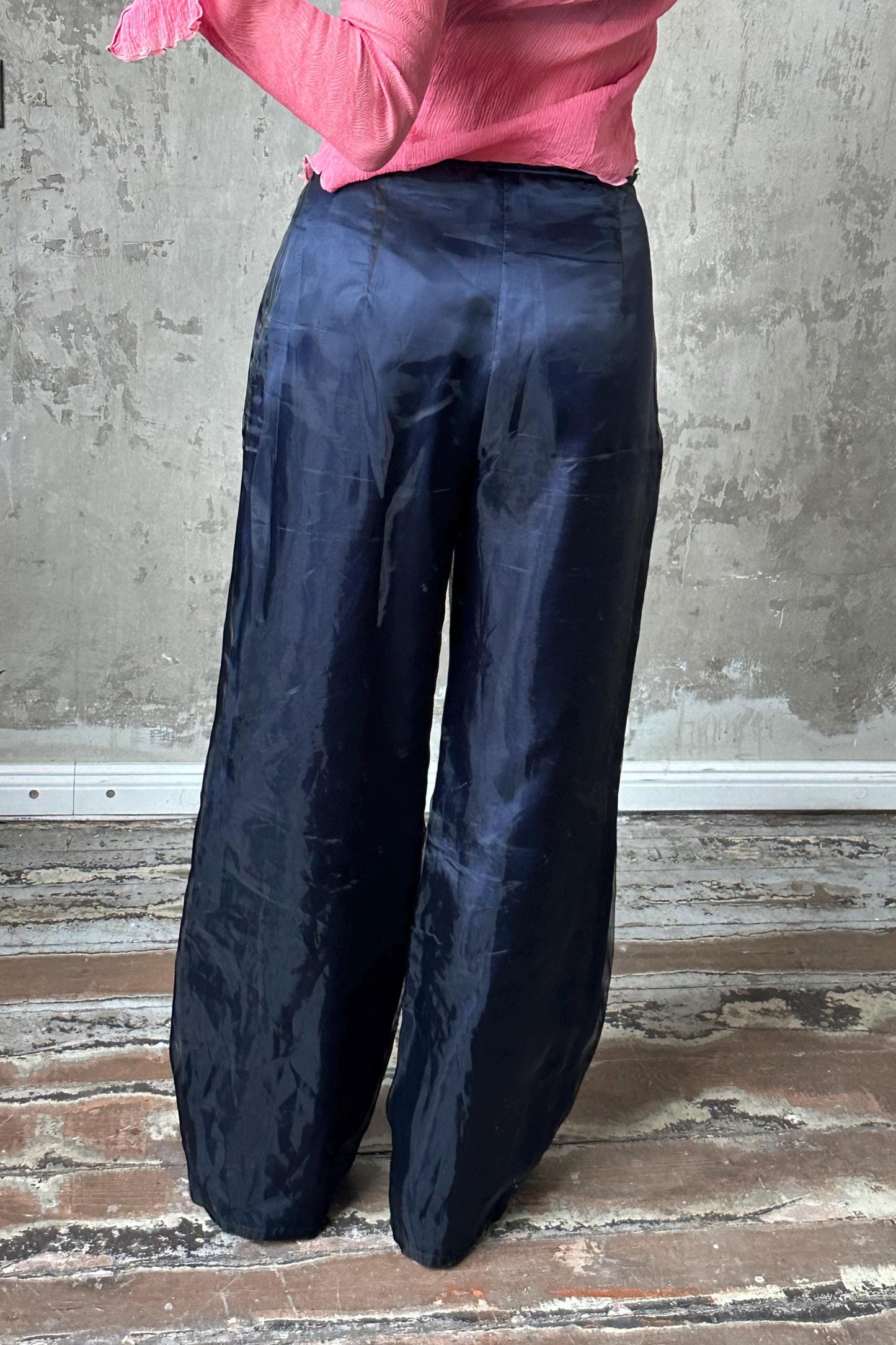 A person wearing a pink top and navy blue wide-leg pants stands in front of a concrete wall.