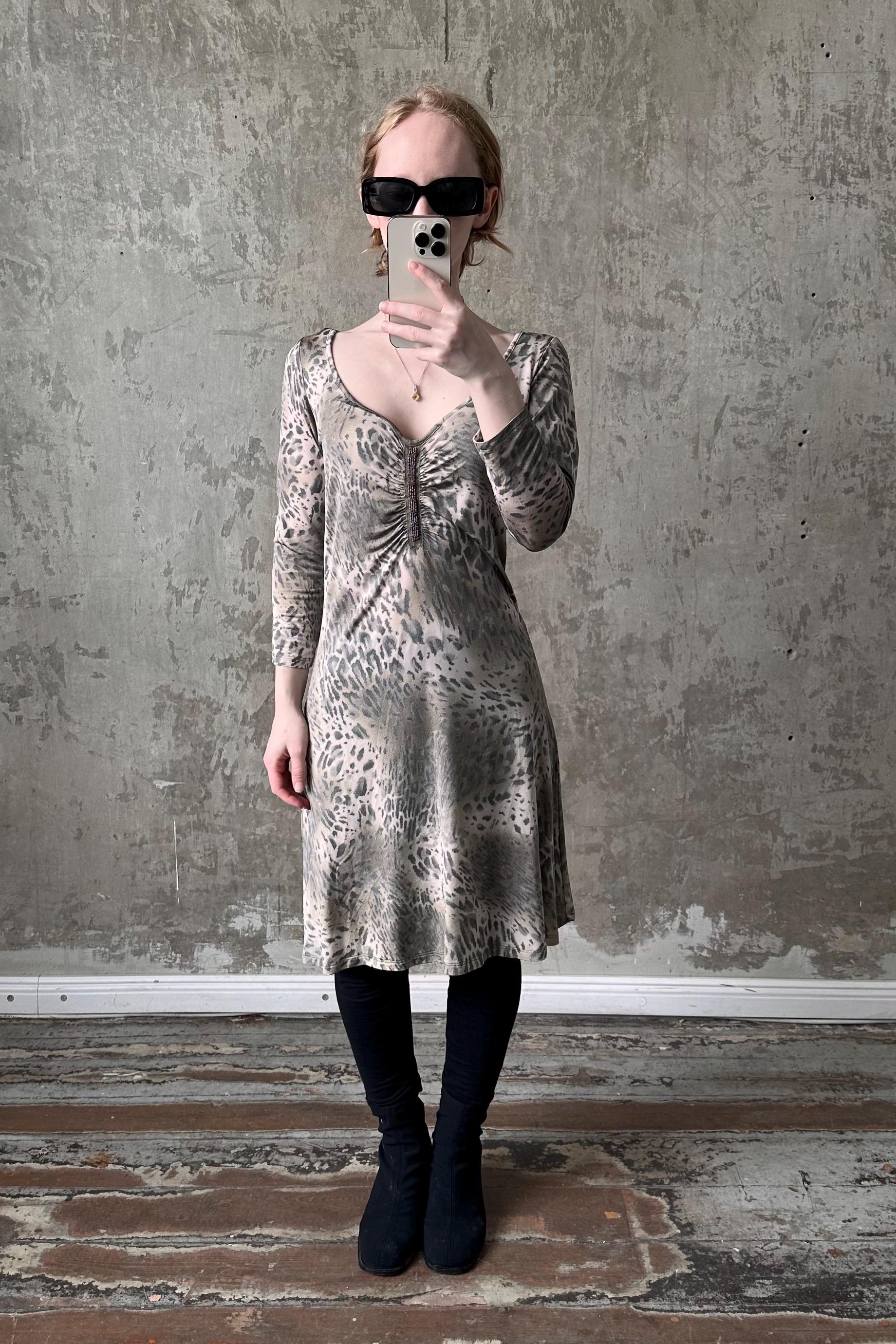 A woman wearing a leopard print dress and sunglasses is taking a selfie with her phone against a concrete wall.