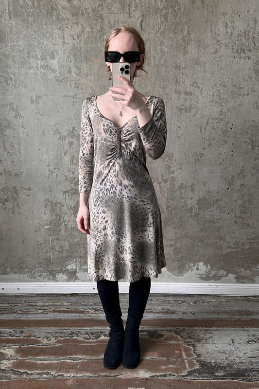 A woman wearing a leopard print dress and sunglasses is taking a selfie with her phone against a concrete wall.