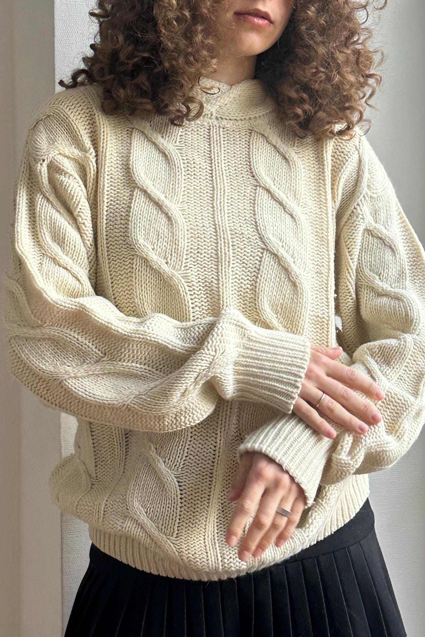 A person with curly hair wearing a cream-colored knitted sweater and a black skirt, posing with their hands crossed.