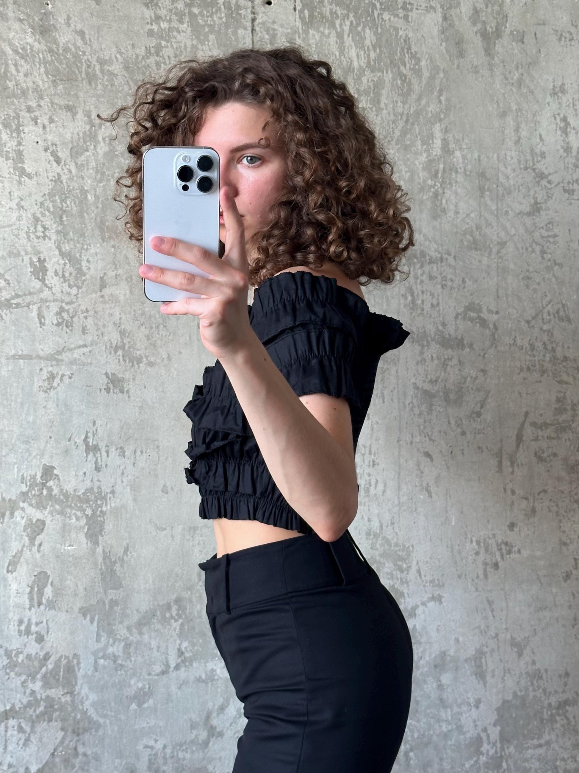 A woman with curly hair is taking a selfie with a smartphone, wearing a black off-the-shoulder top and black pants.
