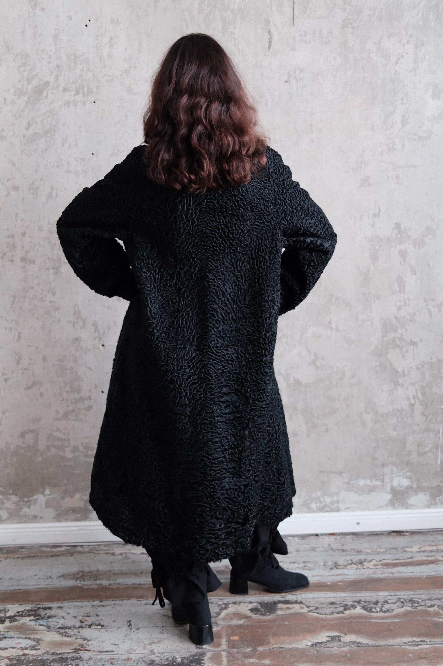 A person wearing a black coat stands in front of a plain white wall.
