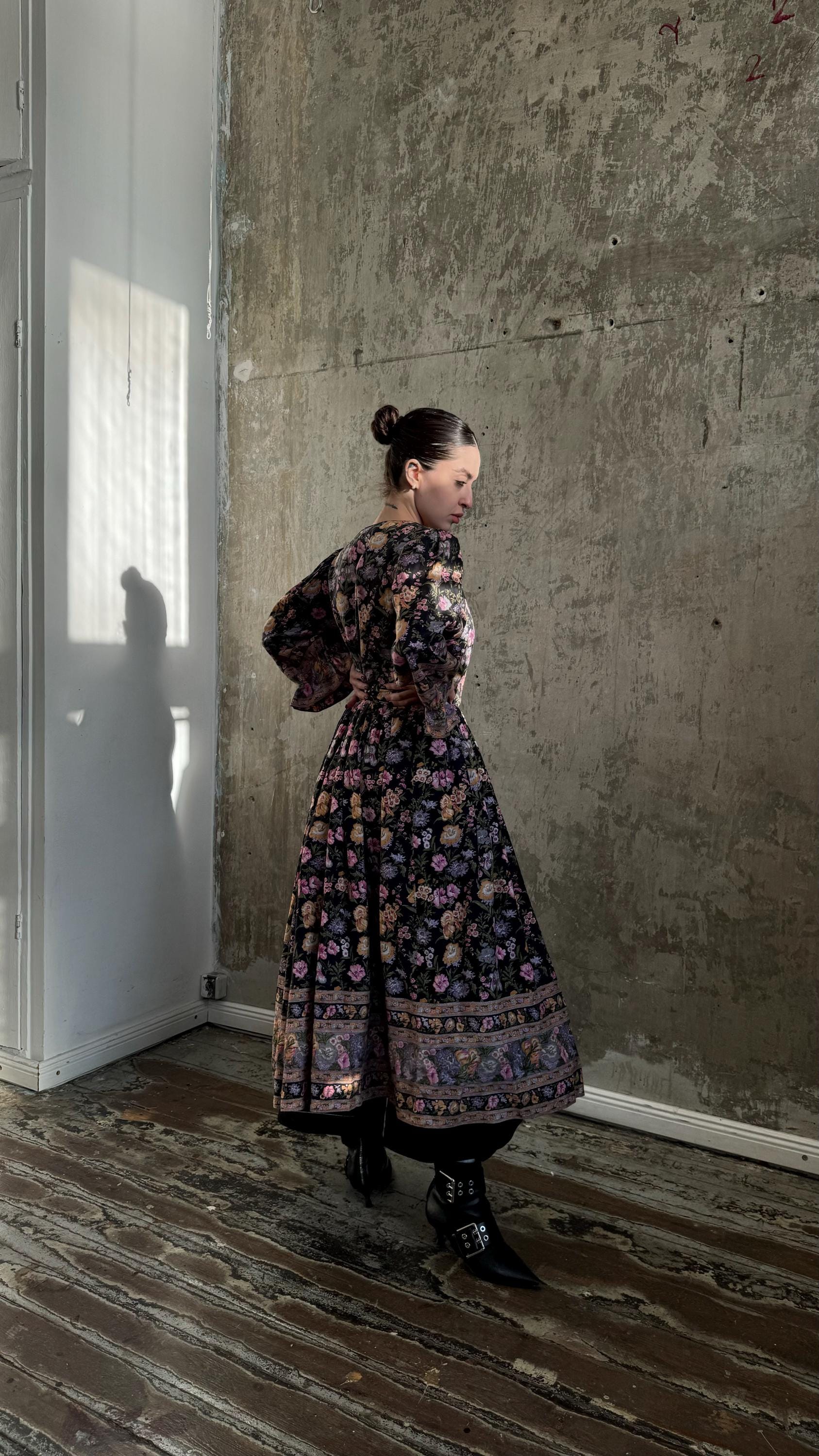 A woman in a long, floral-patterned dress stands in a room with a concrete wall and wooden floor.