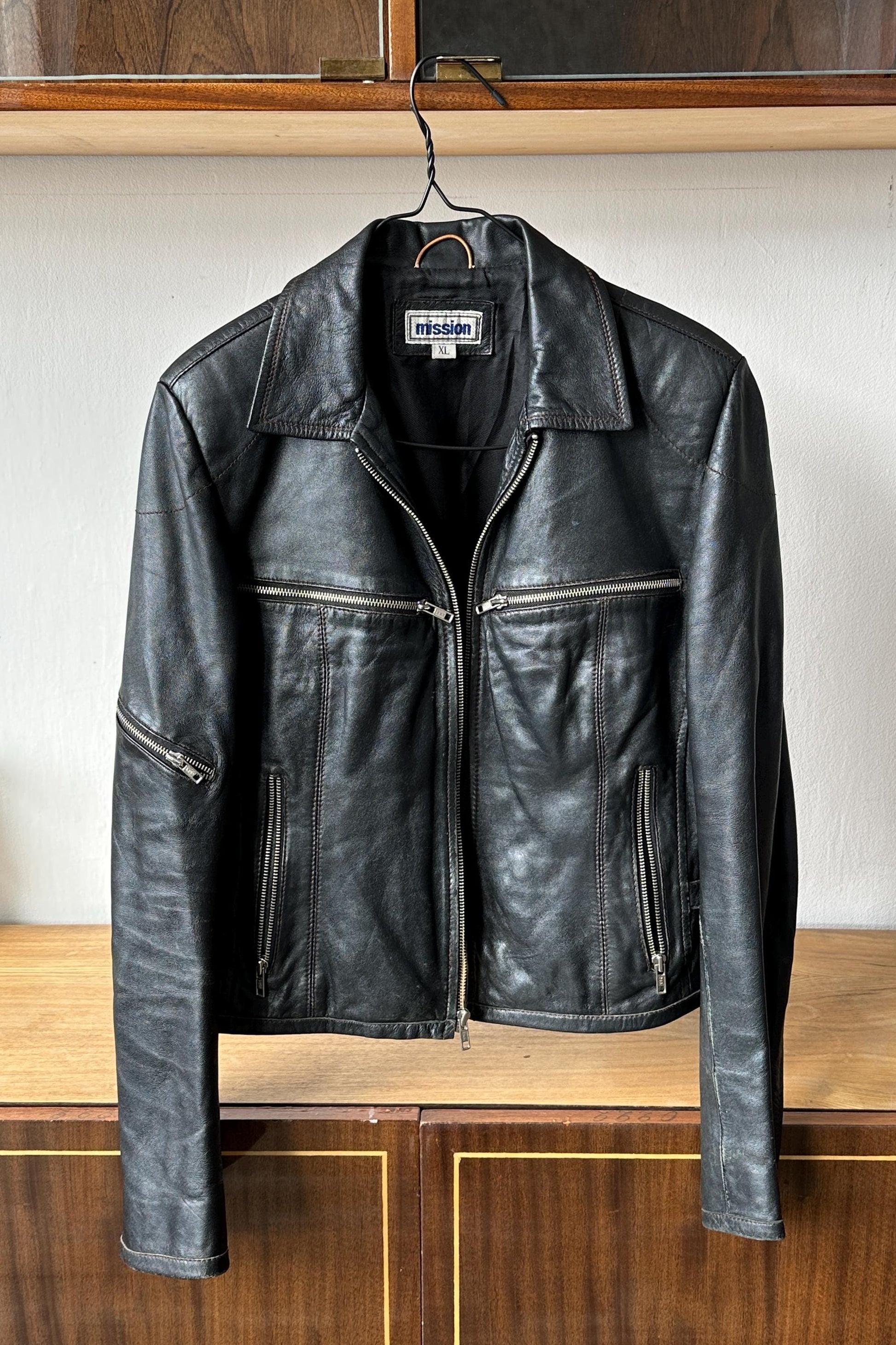 A black leather jacket with a collar and zipper is hanging on a metal hanger against a white wall.