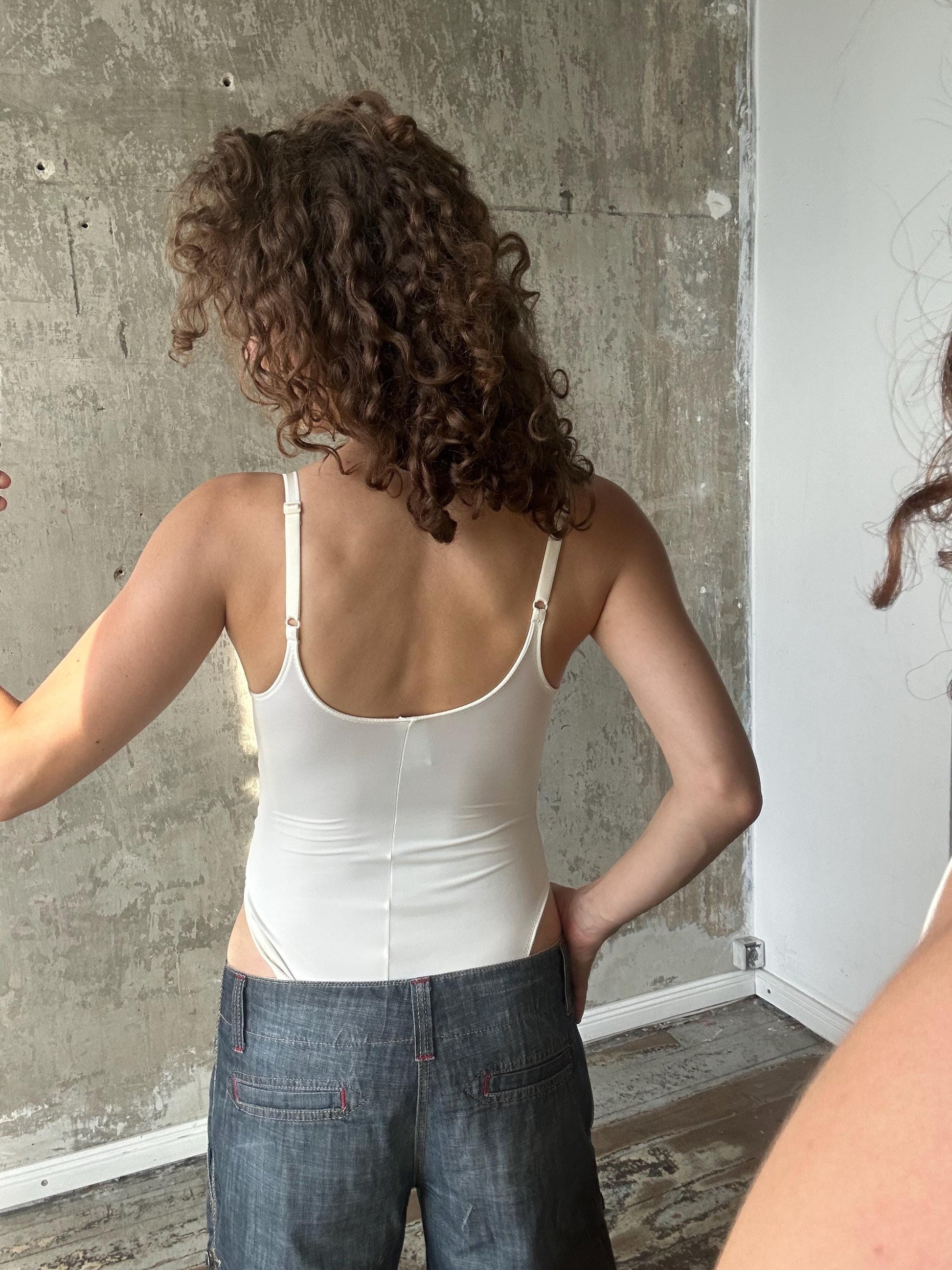 a woman with curly hair wearing a white tank top and blue jeans, standing in front of a concrete wall.