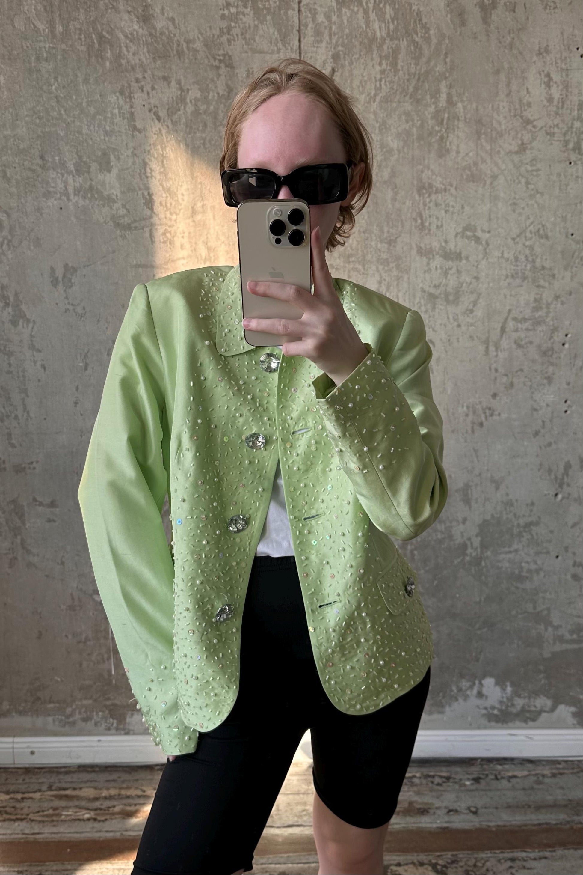 A person wearing a green jacket with sparkly details and sunglasses is taking a selfie with a smartphone.