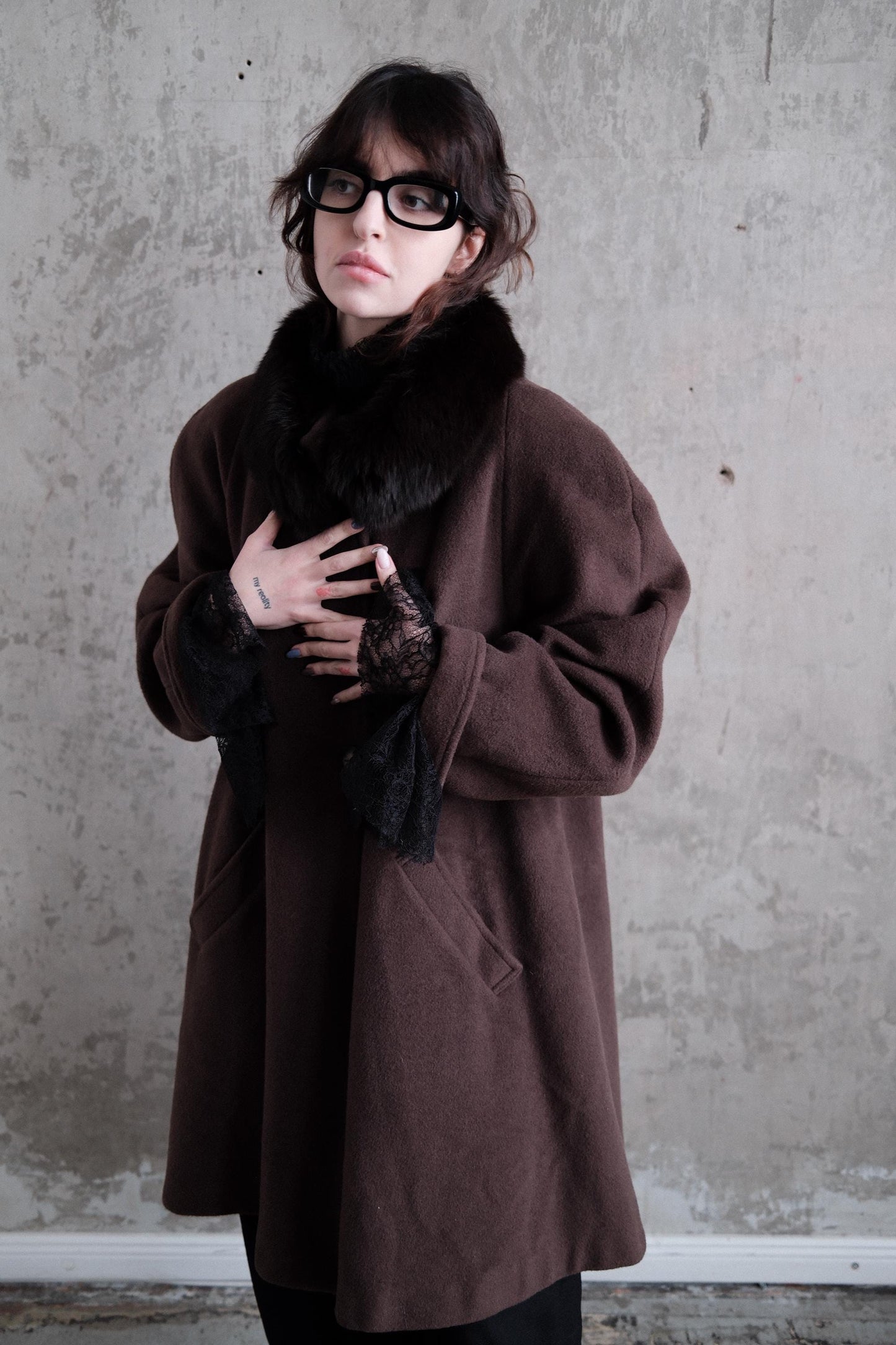 A woman wearing a brown coat and glasses stands against a concrete wall, her gaze directed off to the side.