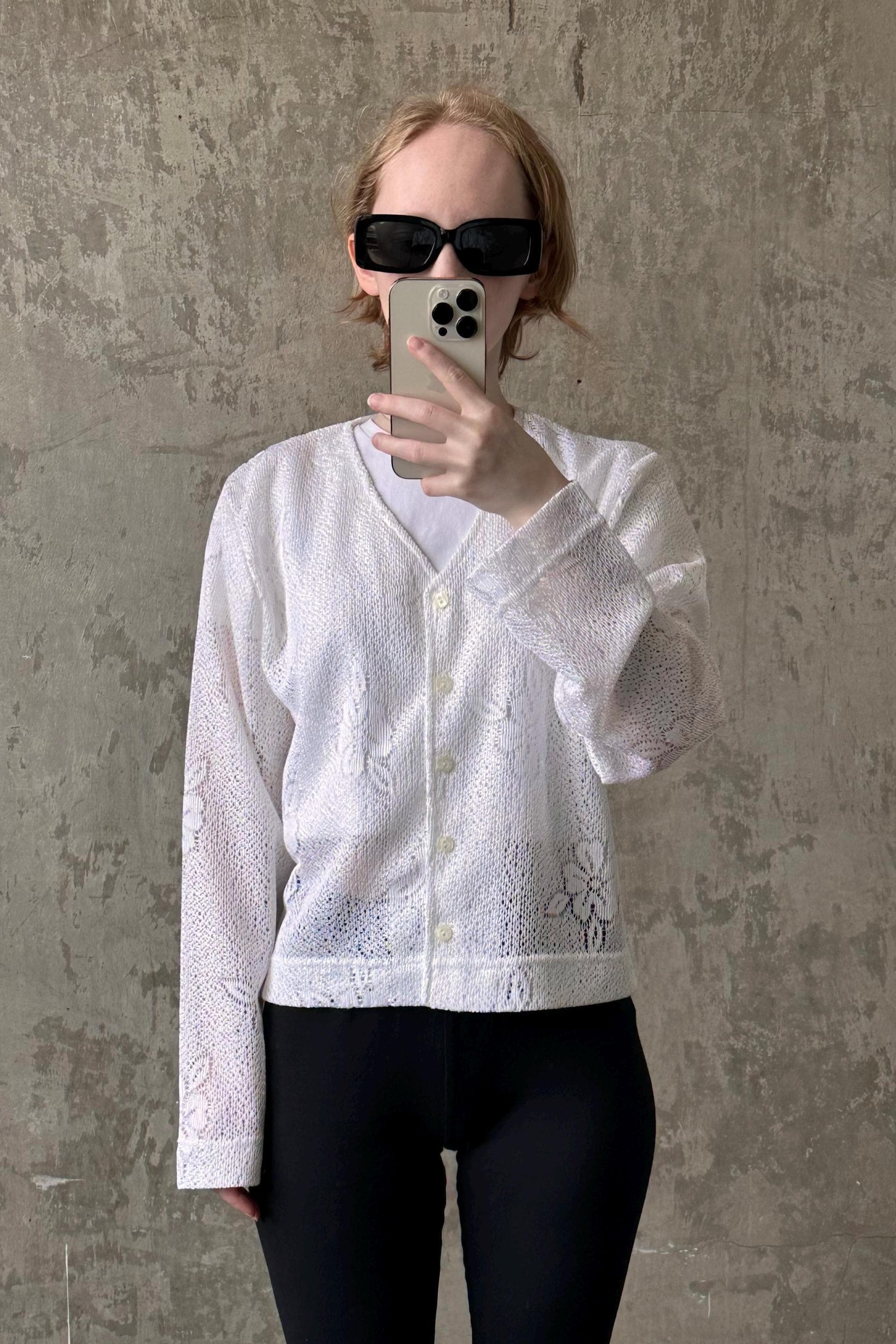 A person wearing a white lace cardigan and sunglasses is taking a selfie with a smartphone.